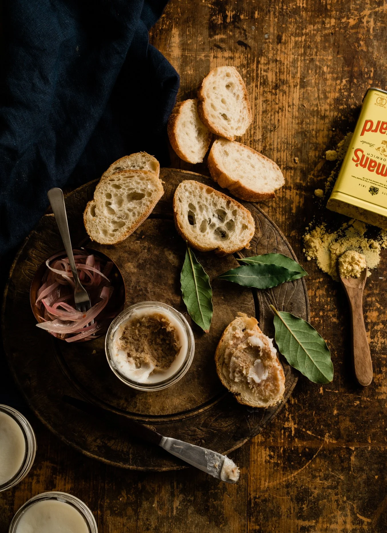 Sliced bread, butter, pickled onions, bay leaves, and butter in a dish on a wooden table, with a black cloth and a box of plain breadcrumbs nearby.