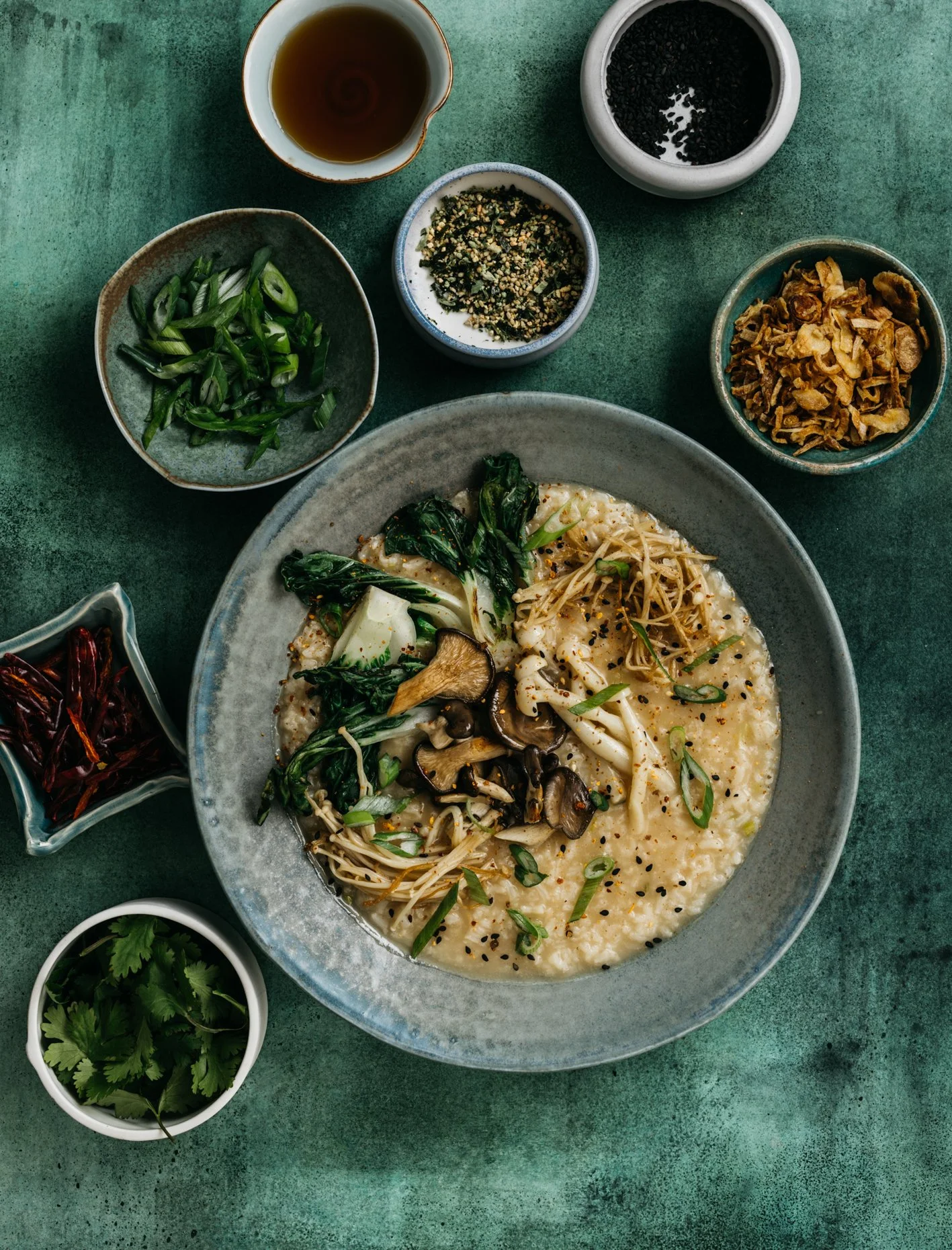 A bowl of creamy ramen topped with vegetables and mushrooms, surrounded by small dishes of herbs, spices, and condiments on a green surface.