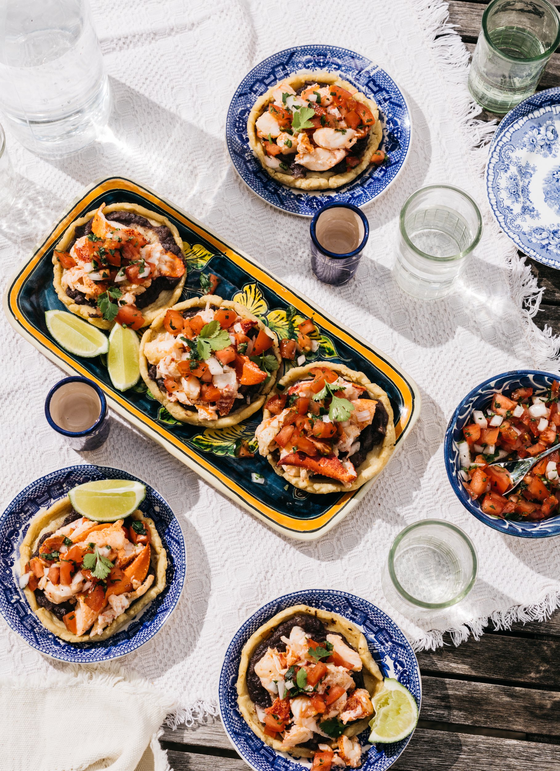 A table set with Mexican tacos topped with shrimp, vegetables, and cilantro, along with lime wedges, glasses of water, and a bowl of fresh salsa. The table features blue and white plates and a white tablecloth.