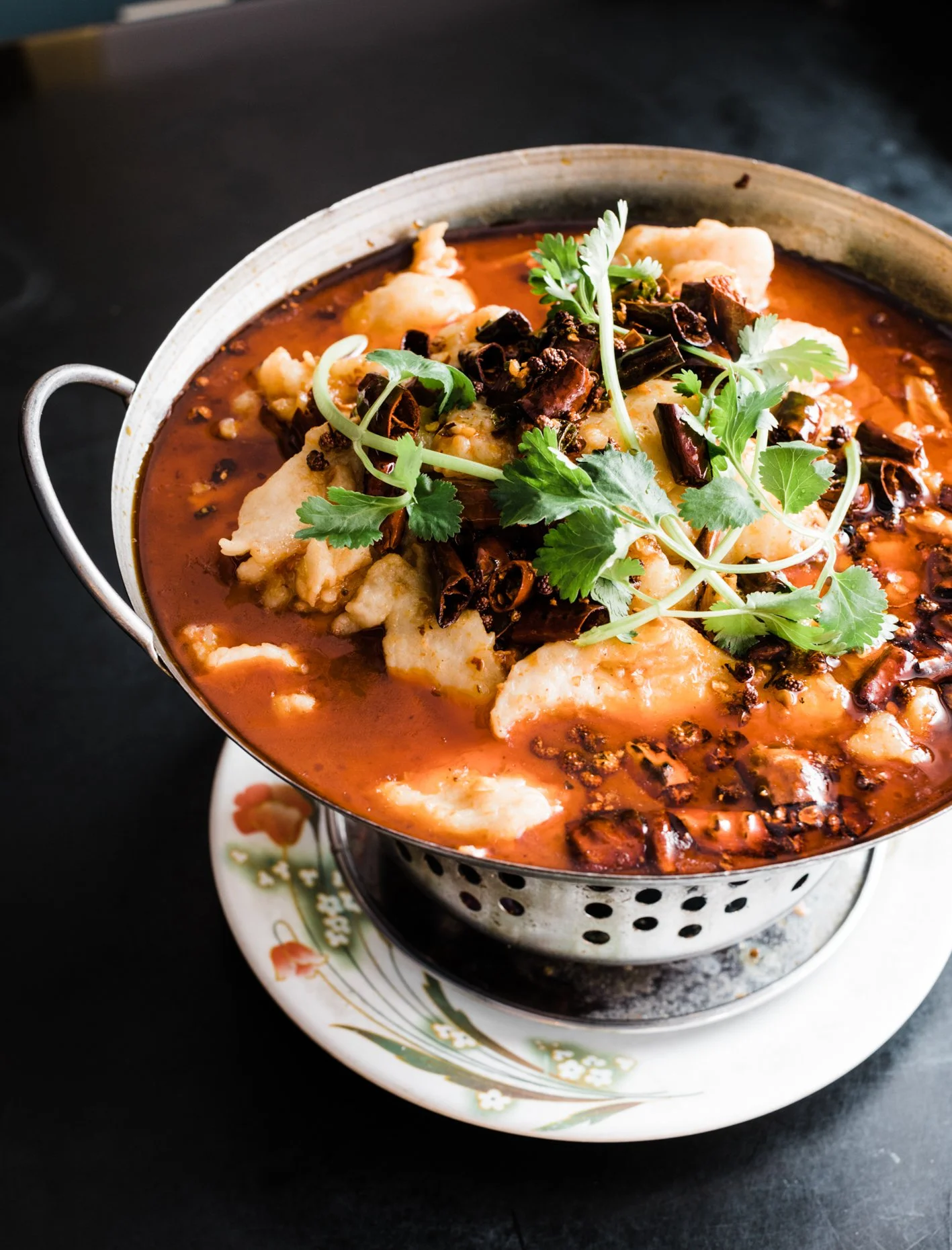 A bowl of spicy Chinese hot pot with chunks of fish, garnished with fresh cilantro and dried chili peppers.