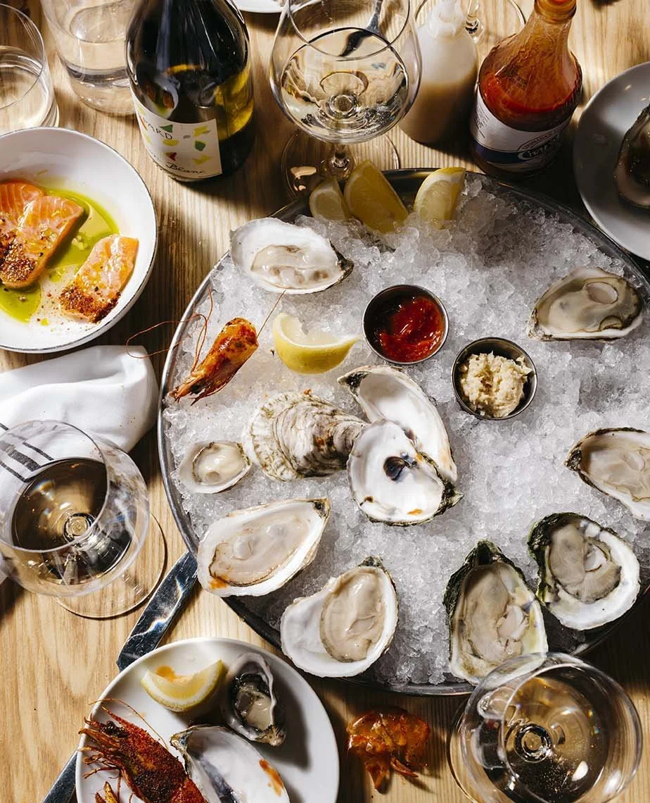 A seafood feast featuring a large platter of fresh oysters on ice with lemon wedges, accompanied by glasses of white wine, and side dishes including salmon, lobster, and dipping sauces on a wooden table.