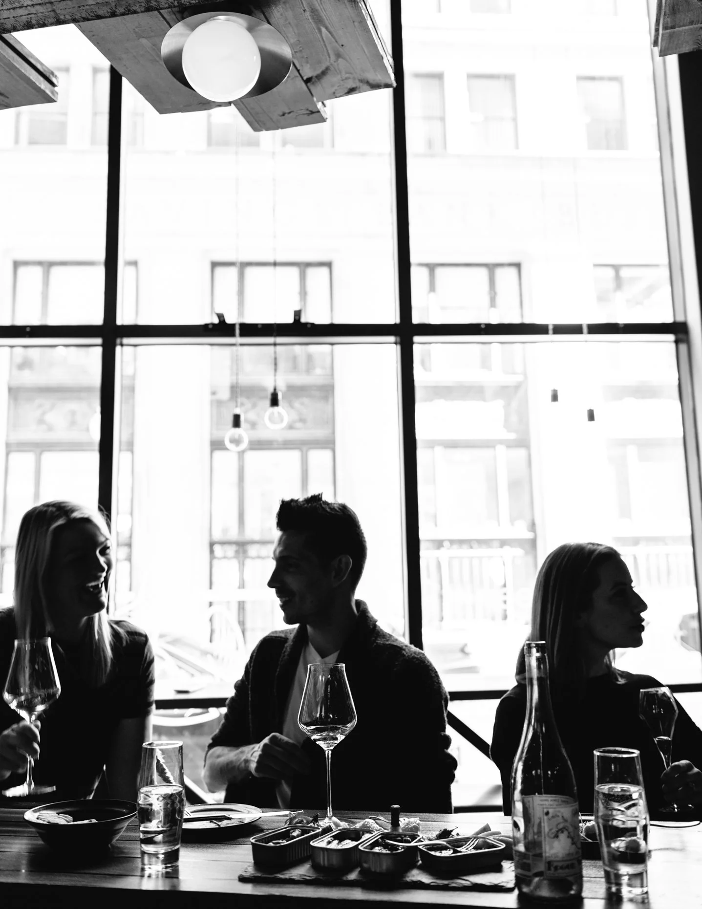 Silhouettes of three people sitting at a table with wine glasses and food, inside a restaurant with large windows and hanging lights.