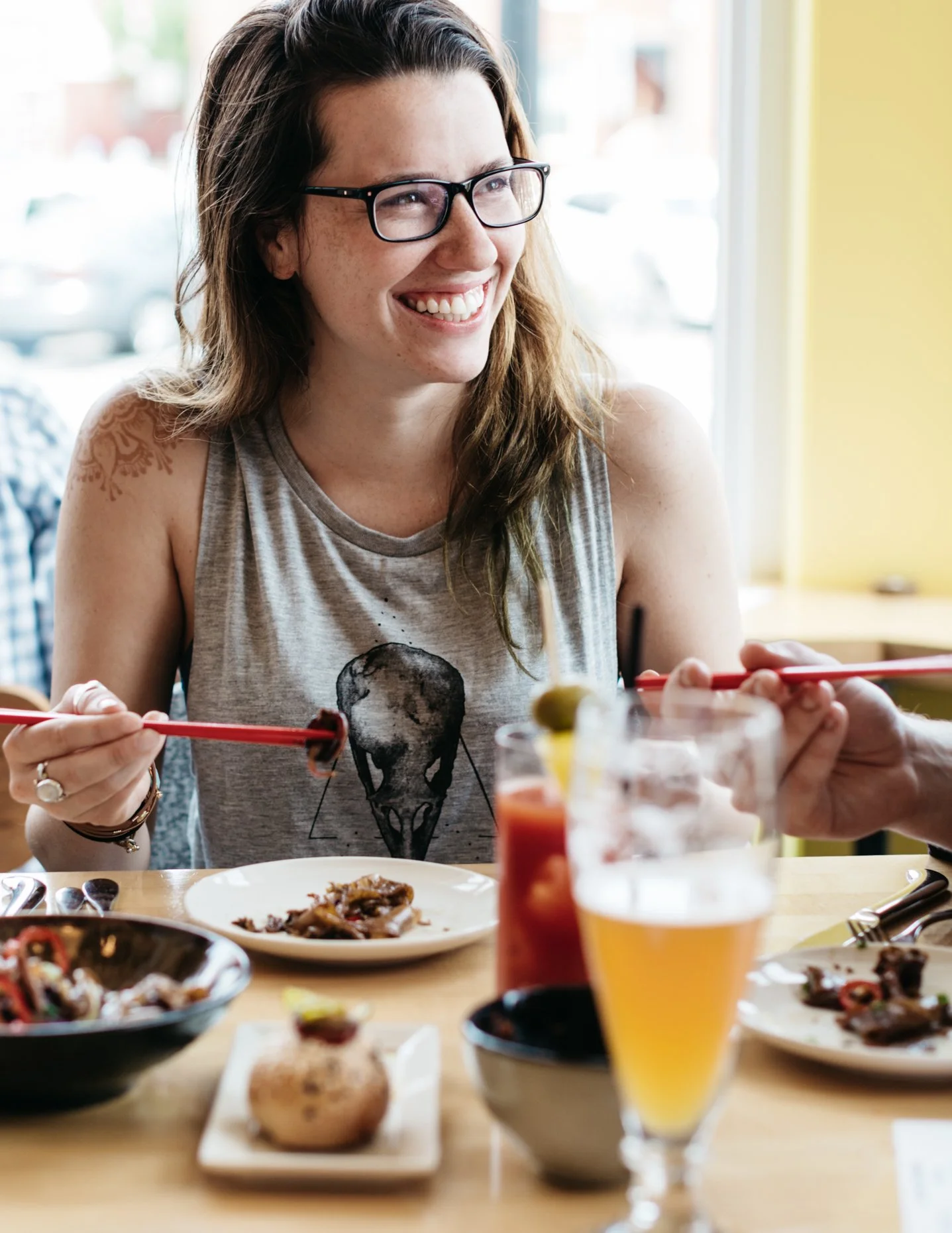 A woman with glasses, long hair, and a sleeveless shirt is smiling while dining at a restaurant. She has a tattoo on her shoulder and is holding chopsticks.