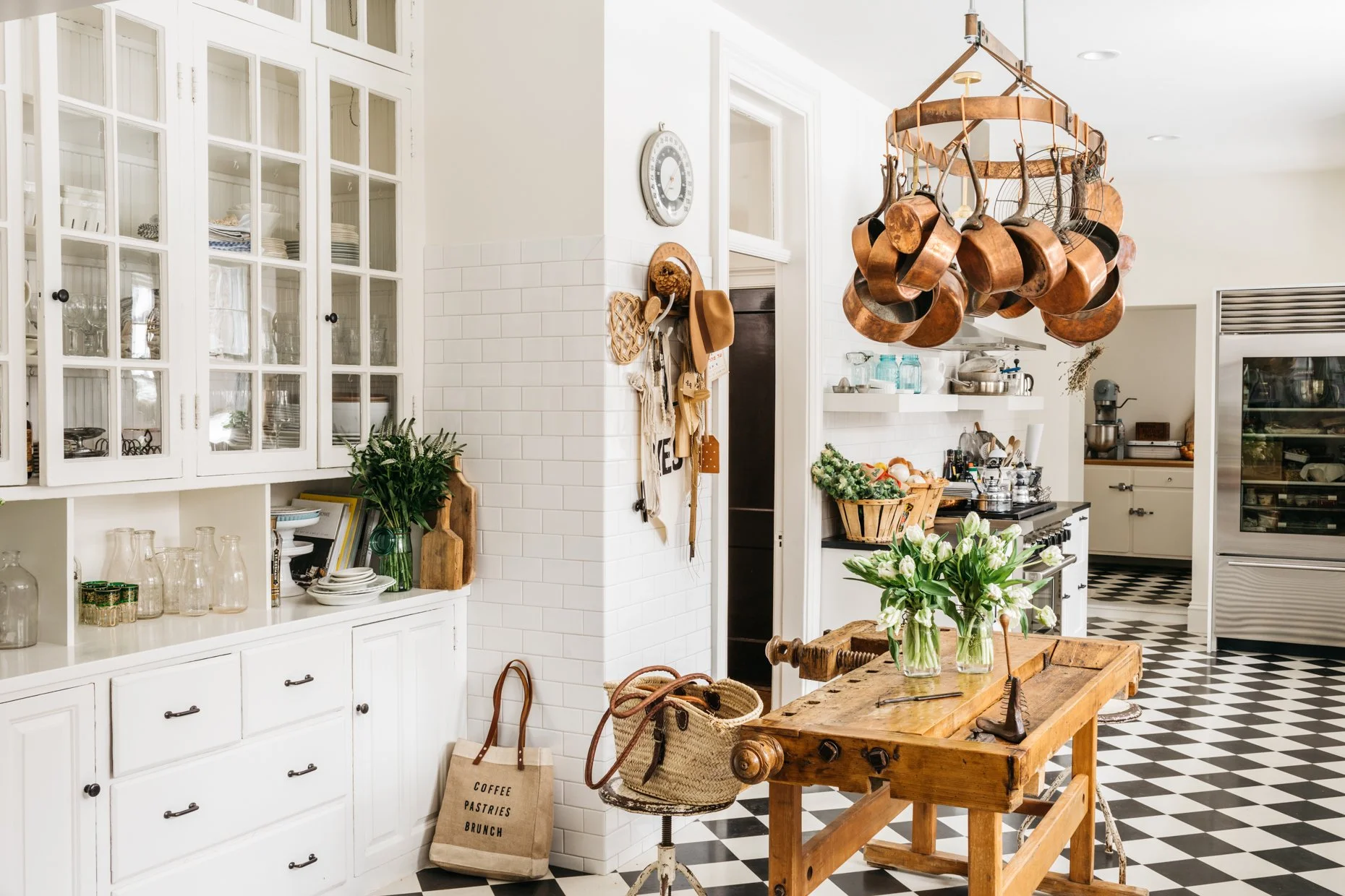 A bright kitchen with white cabinets, a wooden table with flowers, a pot rack with copper pots, black and white checkered floors, and shelves with kitchen utensils.