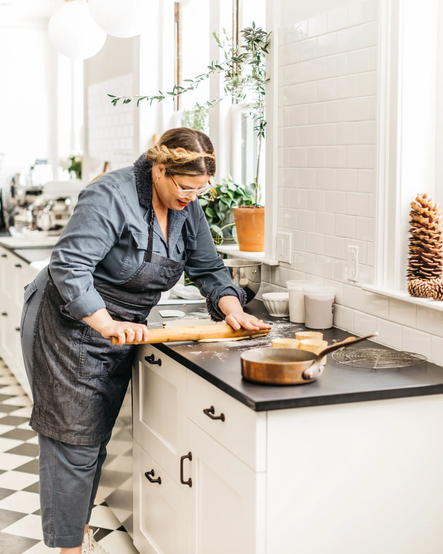 Woman wearing glasses and a black apron rolling dough with a rolling pin on a kitchen counter.