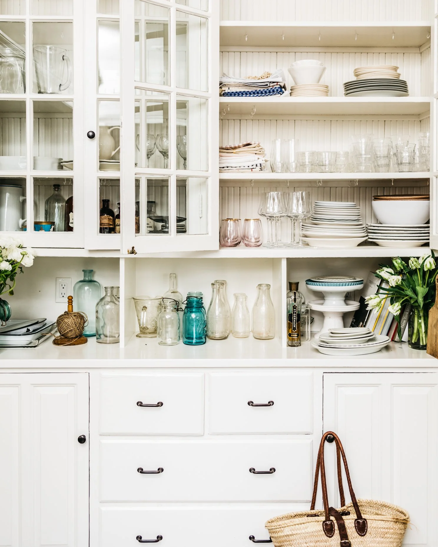 White kitchen cabinet filled with glassware, dishes, and decorative jars, with a woven basket and tote bag on the floor.