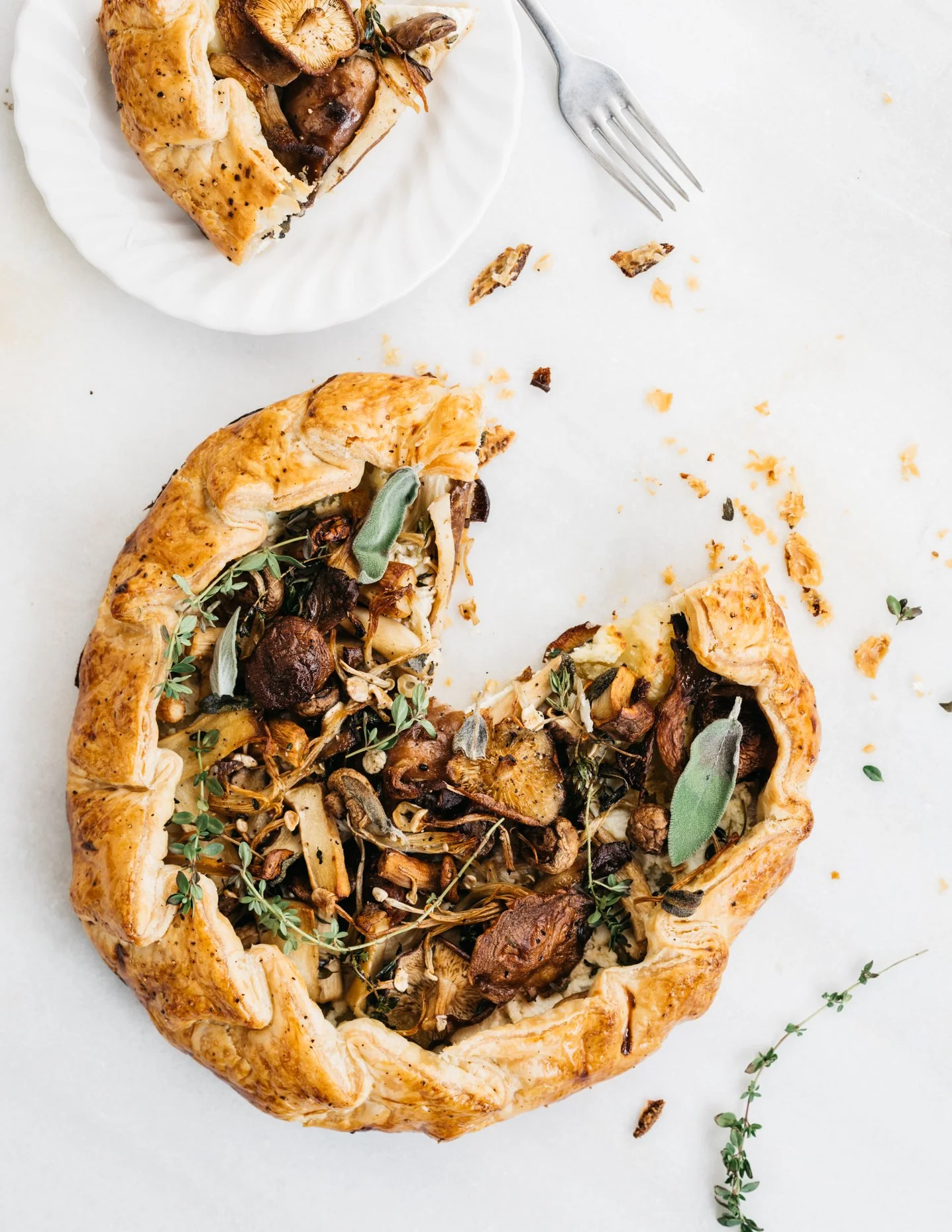 Mushroom and herb galette with chopped herbs and a bite taken out of it, on a white surface with a small white plate and a fork nearby.