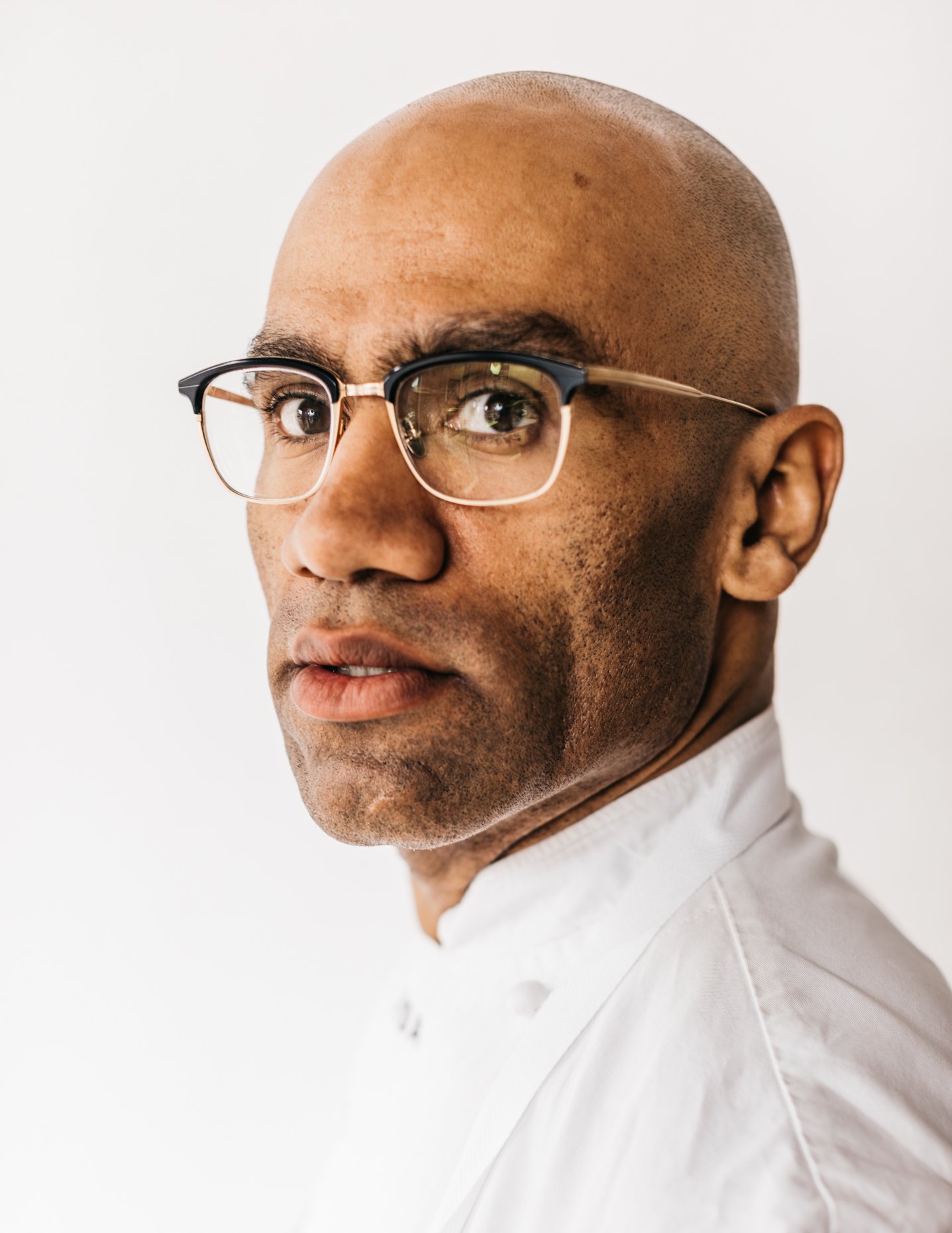A close-up of a man with a shaved head and glasses, wearing a white shirt, against a plain white background.