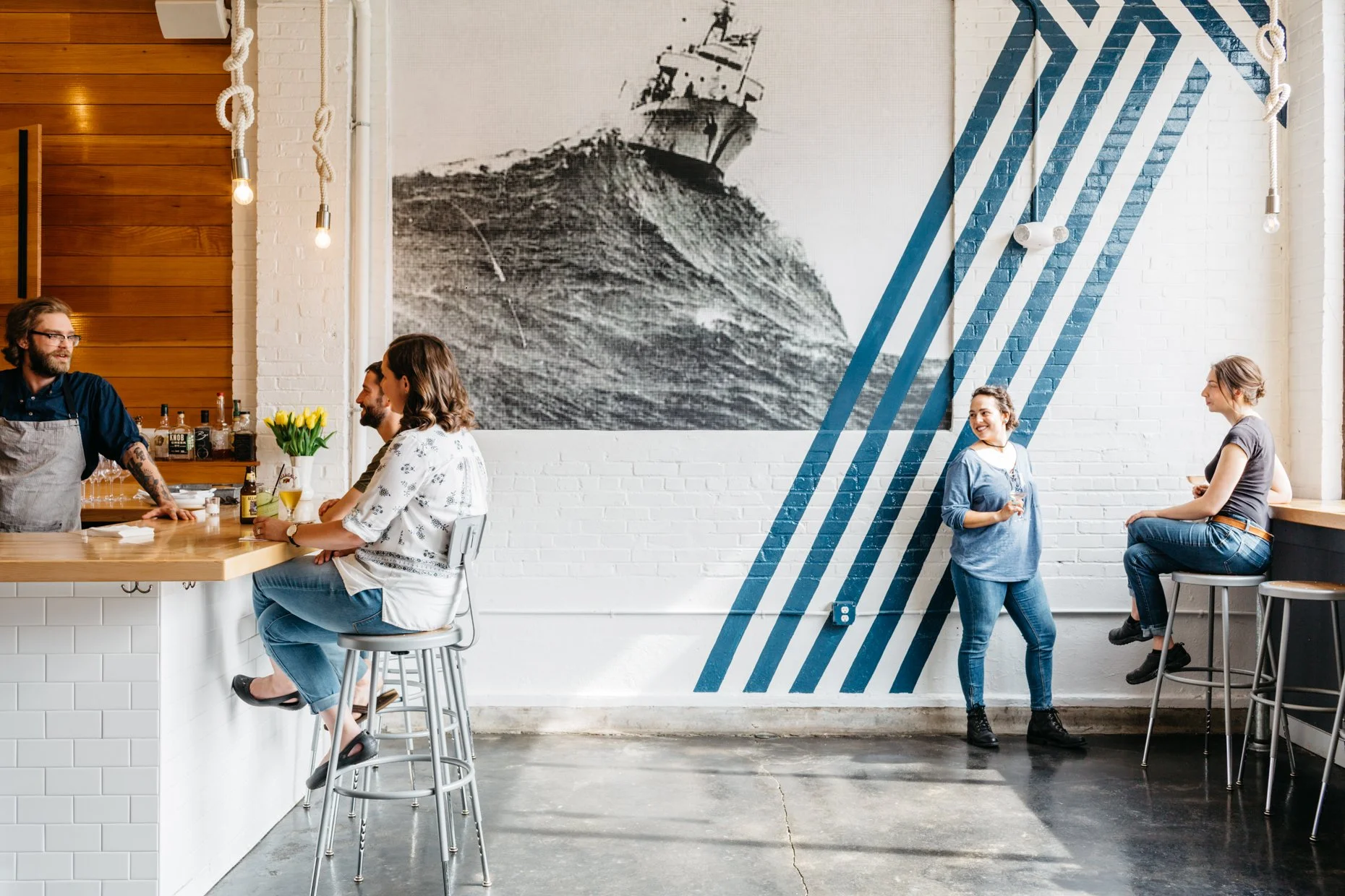 People socializing inside a modern cafe with a large black-and-white mural of a ship on the wall and blue striped decor.