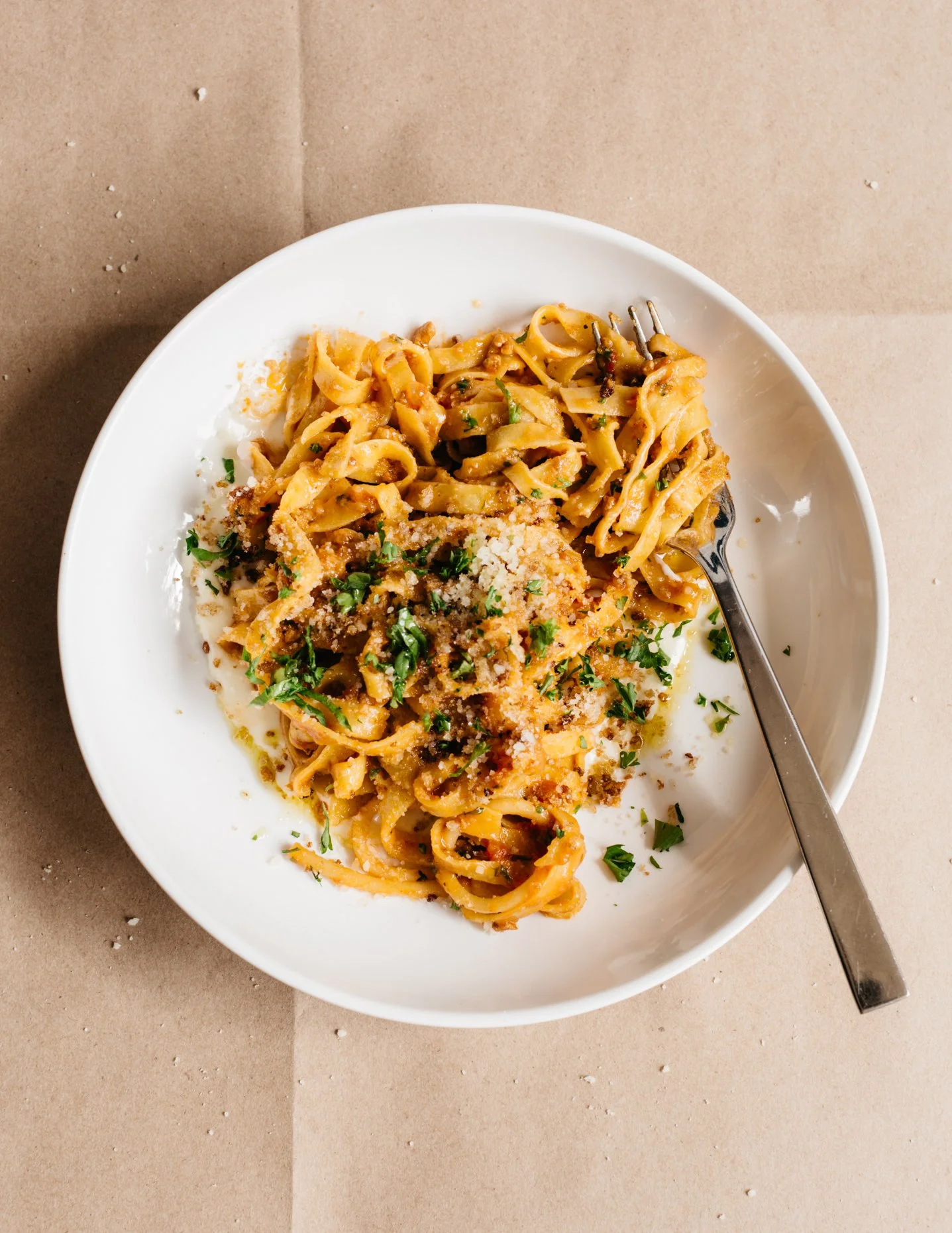 A white bowl of fettuccine pasta with tomato sauce, grated cheese, and chopped parsley, with a fork resting inside the bowl. The bowl is on a brown paper surface.