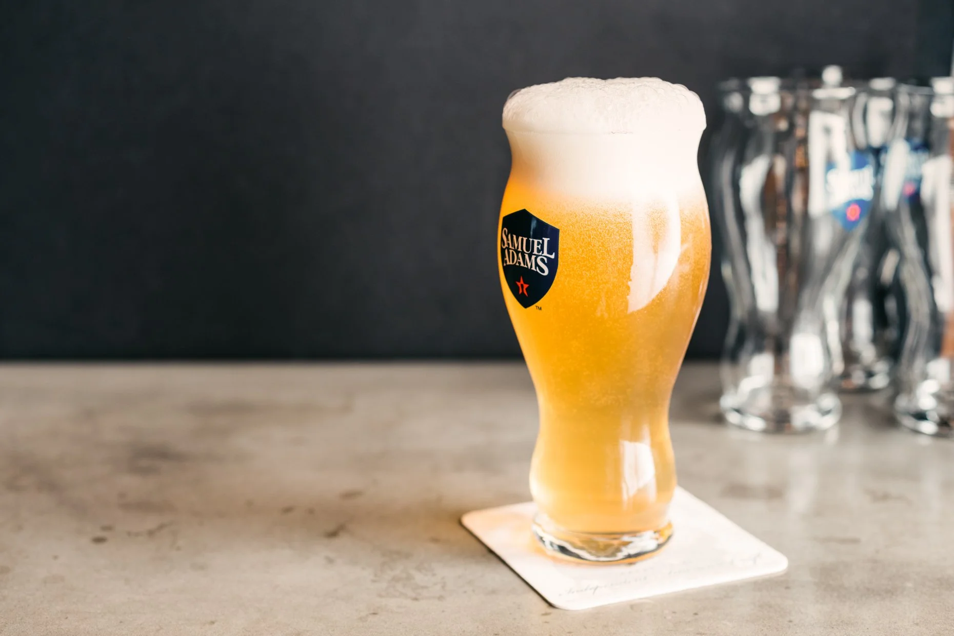 A tall, curved glass of Samuel Adams beer with a thick foamy head on a white coaster, on a wooden surface with a dark background.