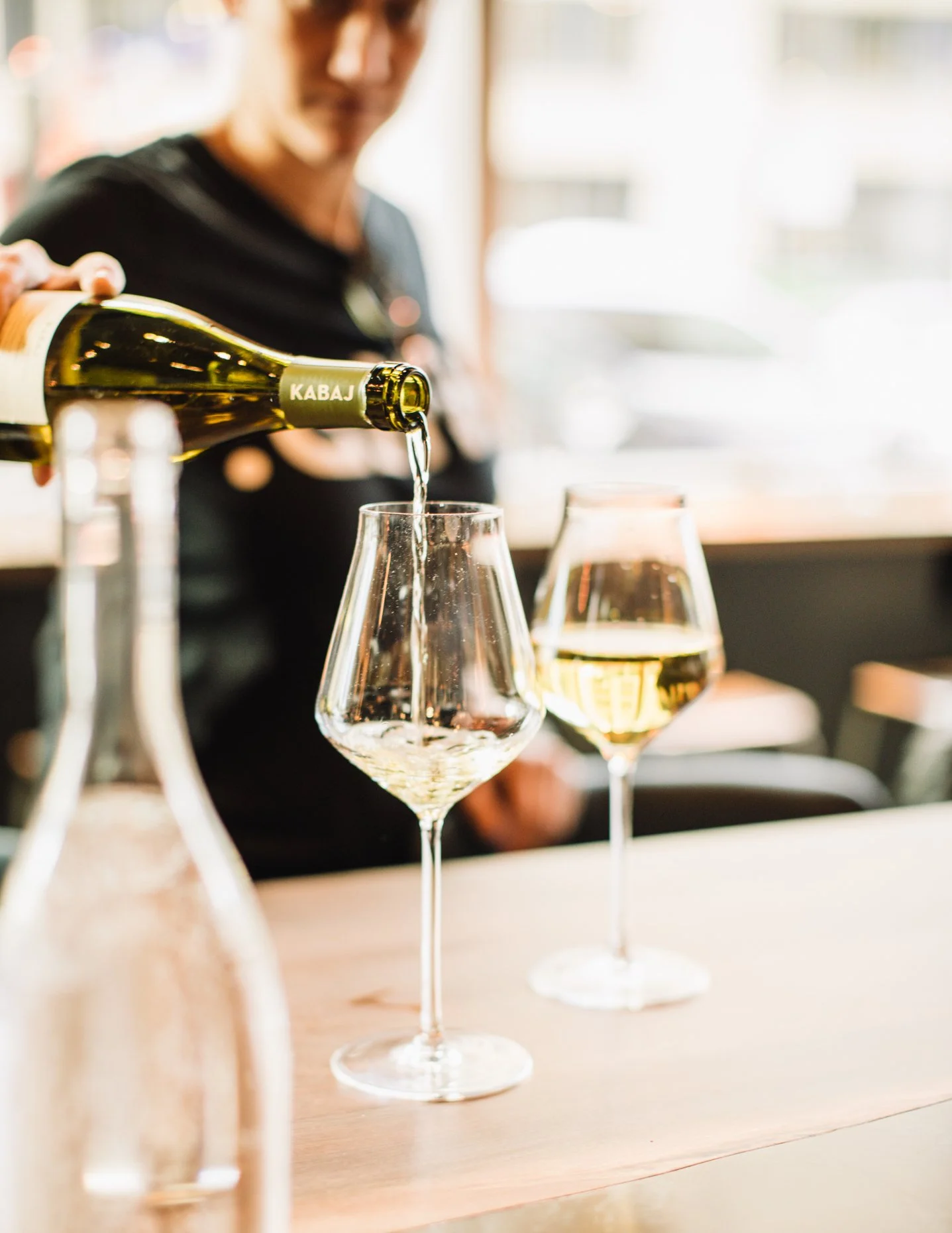 A person pouring white wine into a wine glass at a table with two glasses of wine, one being filled and one already filled, in a bright indoor setting.