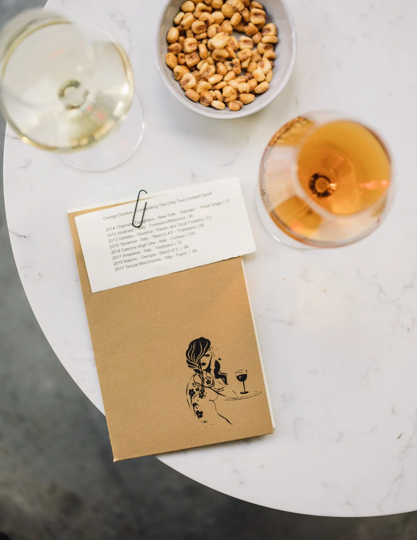 A white marble table with a glass of white wine, a glass of rosé wine, a bowl of chickpeas, and a brown paper menu featuring a woman holding a glass of wine, with a list of wines from various years and countries attached with a paper clip.