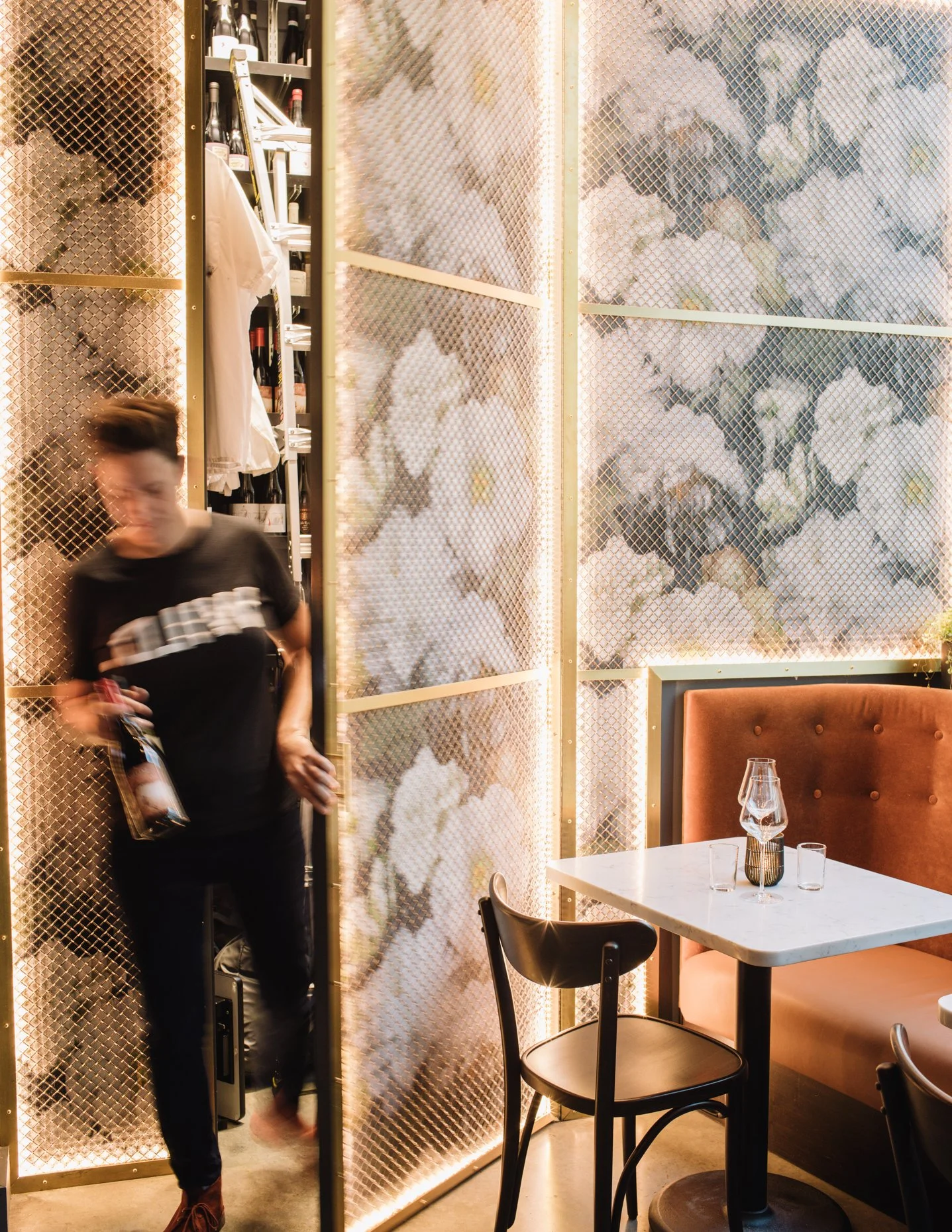 A blurred person in a black t-shirt exiting a restaurant or cafe, with a marble table, empty glasses, and orange seating in the background, illuminated by warm lighting.