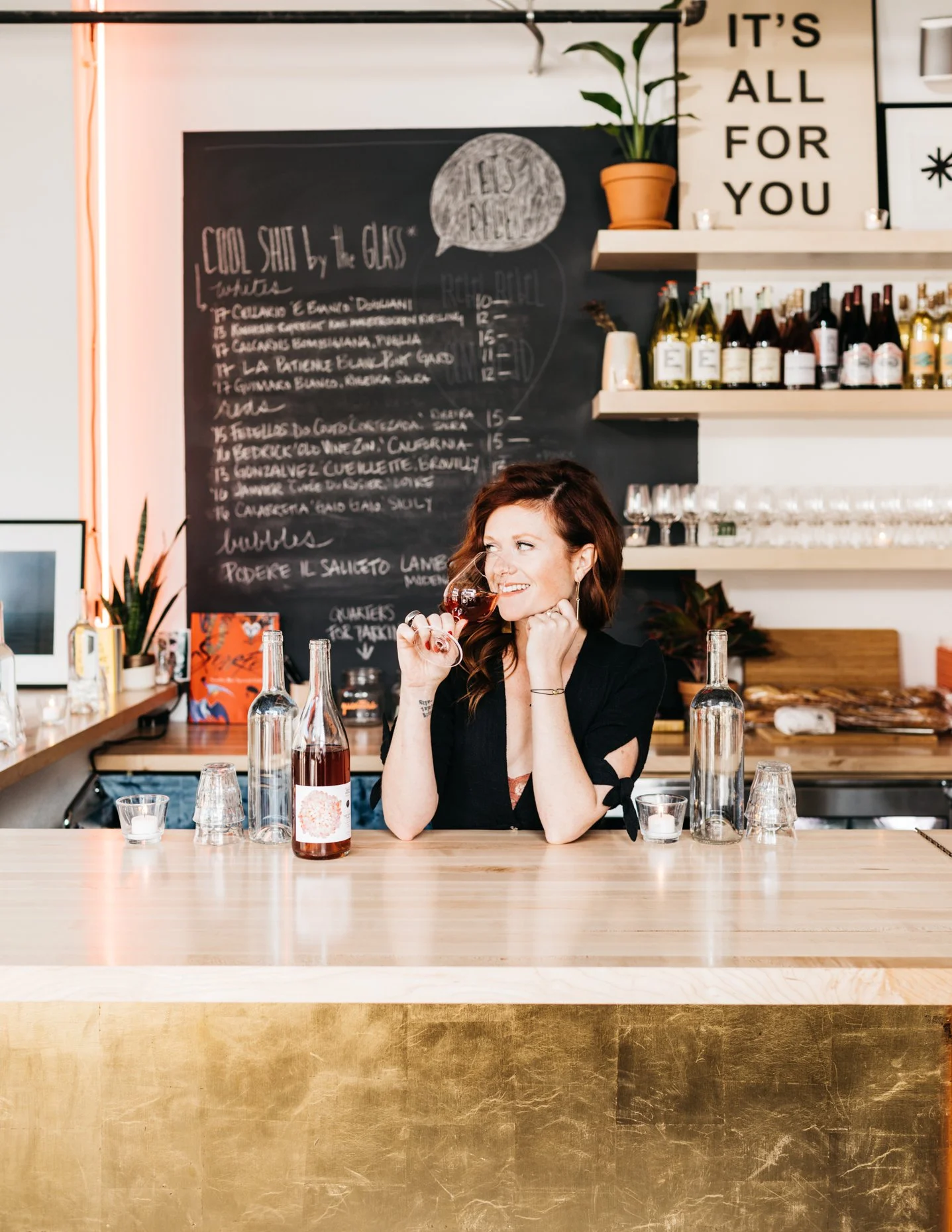 A woman sitting at a bar, holding a glass of red wine and smiling. The bar has bottles of wine and glasses behind her, and a chalkboard menu on the wall. There's a plant and a sign with the text 'IT'S ALL FOR YOU' on the shelves.