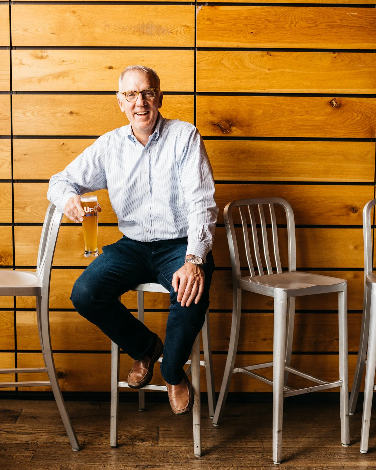 A man sitting on a stool holding a glass of beer, smiling, with a wooden wall background and empty chairs around him.