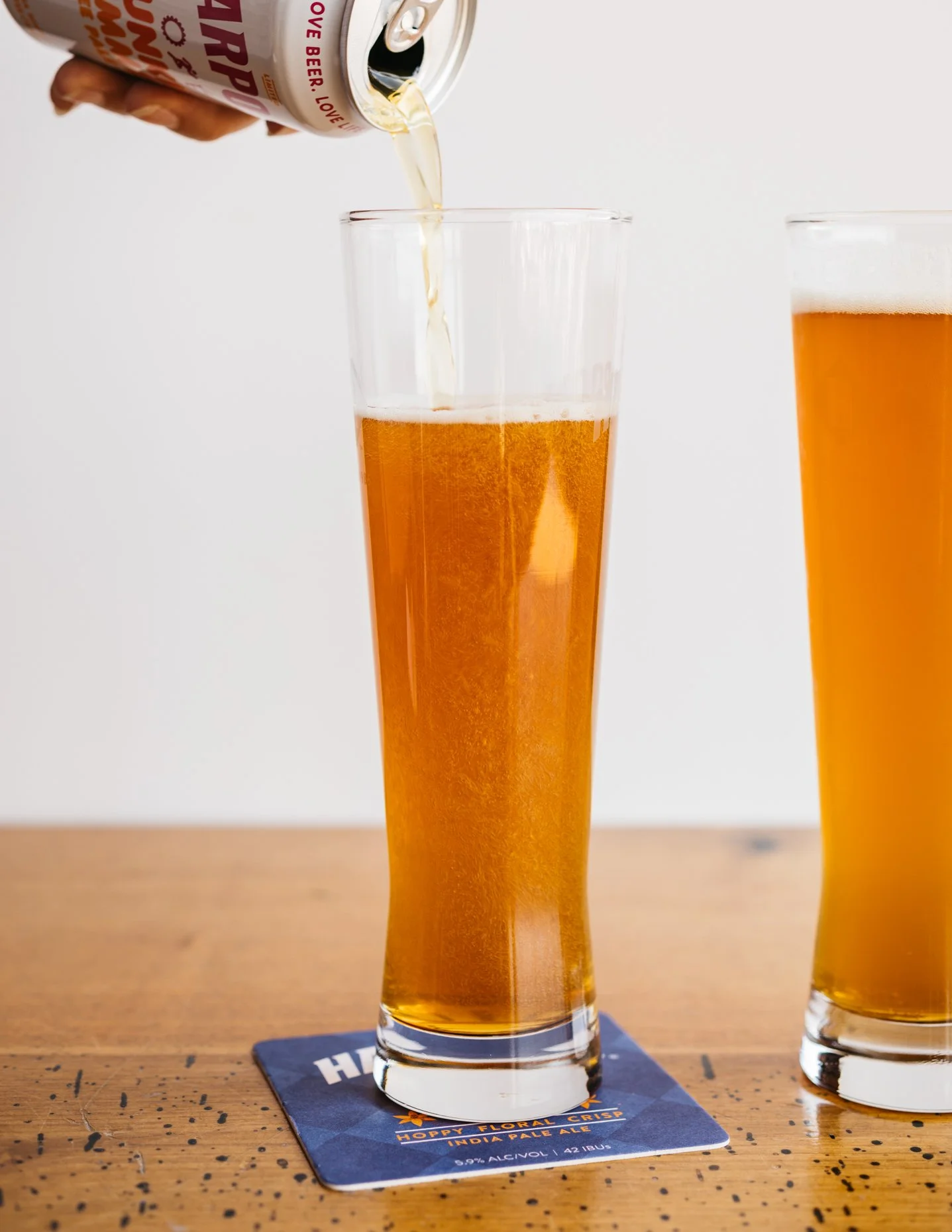 A hand pouring beer from a can into a tall glass filled with amber-colored beer on a wooden table, with another filled glass beside it.