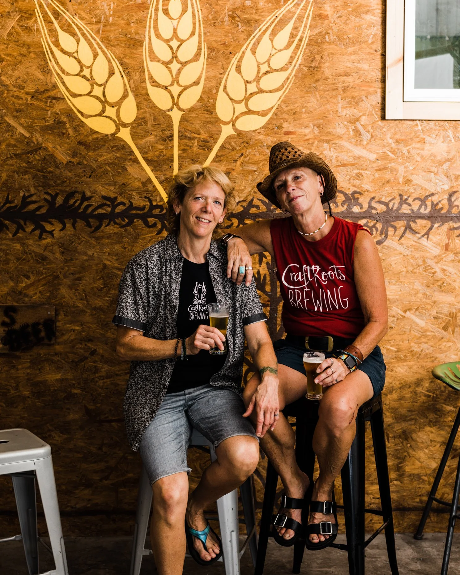 Two women sitting on barstools with drinks in hand, smiling and posing together inside a brewery with wooden wall decor and a wheat design painting behind them.