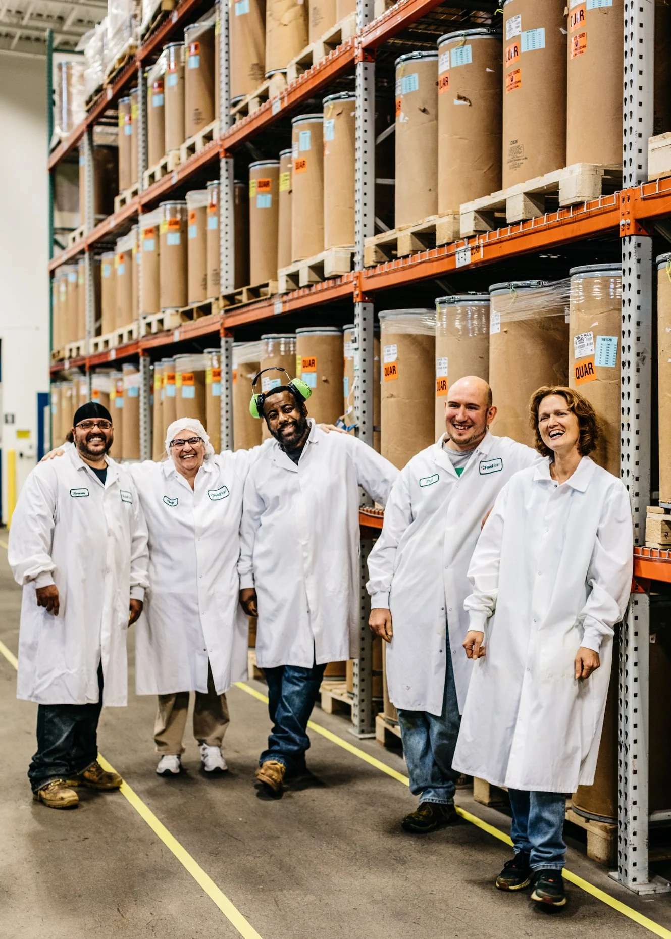 Group of five people in white lab coats smiling and standing in a warehouse aisle with large brown paper and plastic containers on metal shelves behind them.