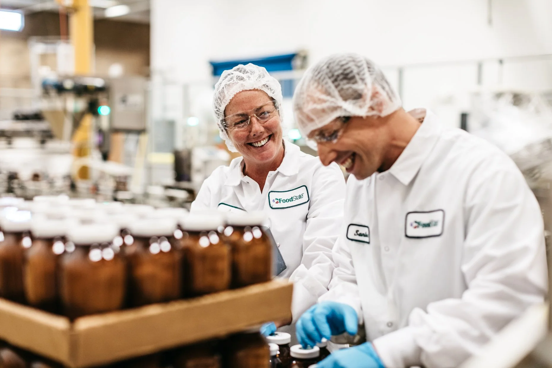 Two smiling workers in a factory wearing white coats, hairnets, and gloves, inspecting bottles on a production line.