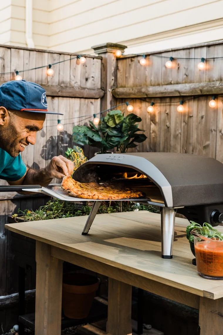 A man wearing a blue cap and teal shirt takes a slice of pizza from a pizza oven on a patio decorated with string lights and plants.