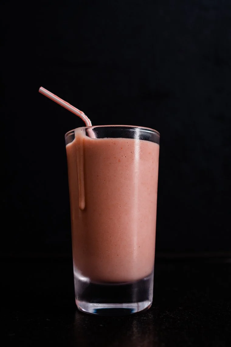 A tall glass of pink strawberry smoothie with a red and white striped straw against a black background.