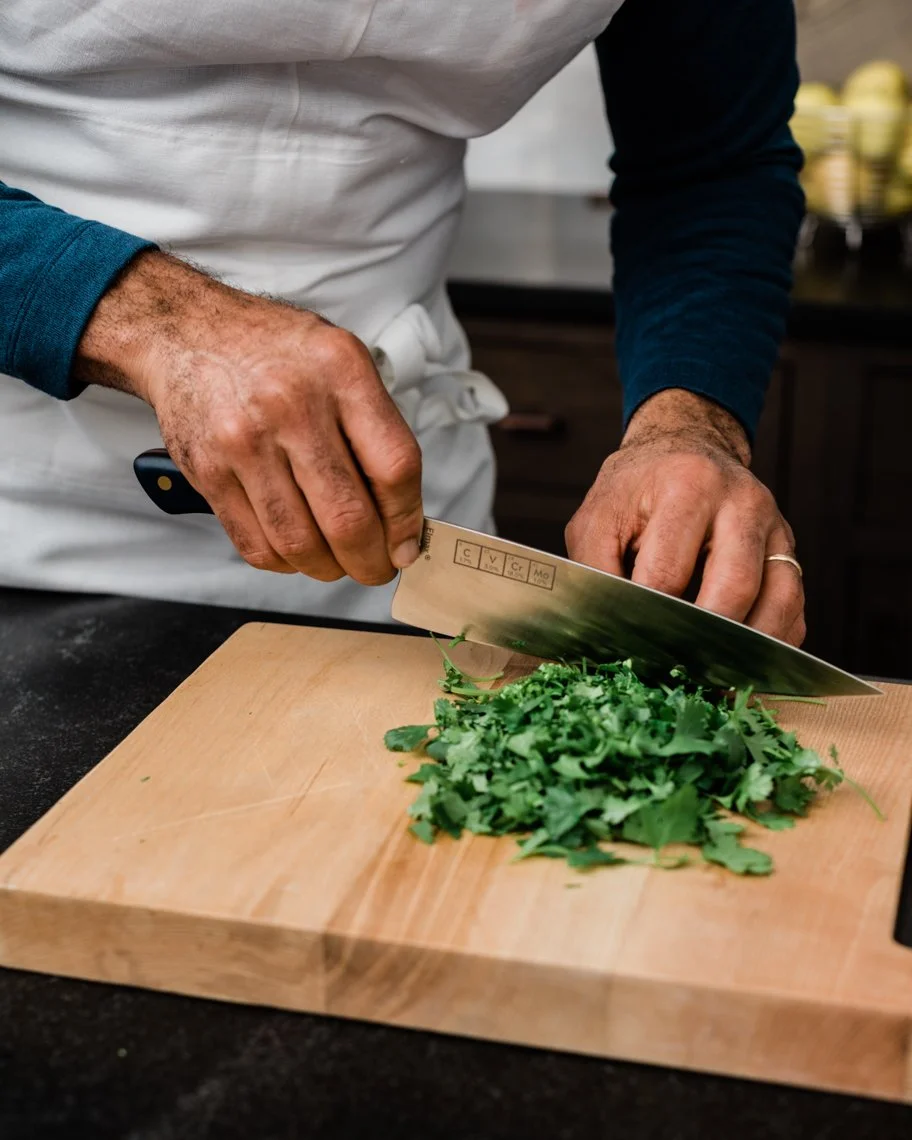 A person chopping fresh green herbs on a wooden cutting board in a kitchen.