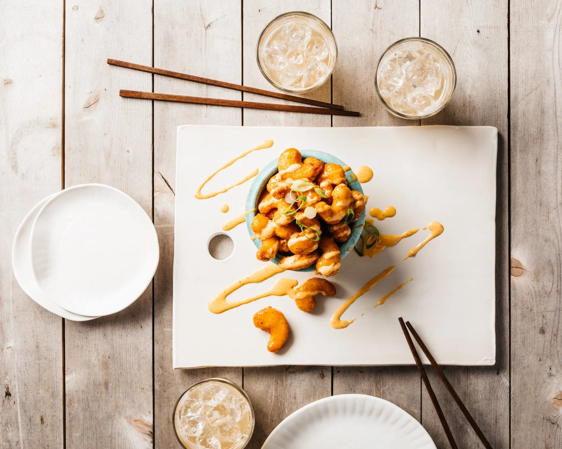 A plate of Asian-style plant-based shrimp with sauce and garnish, served on a white square plate on a wooden table with glasses of iced tea, empty bowls, and chopsticks.