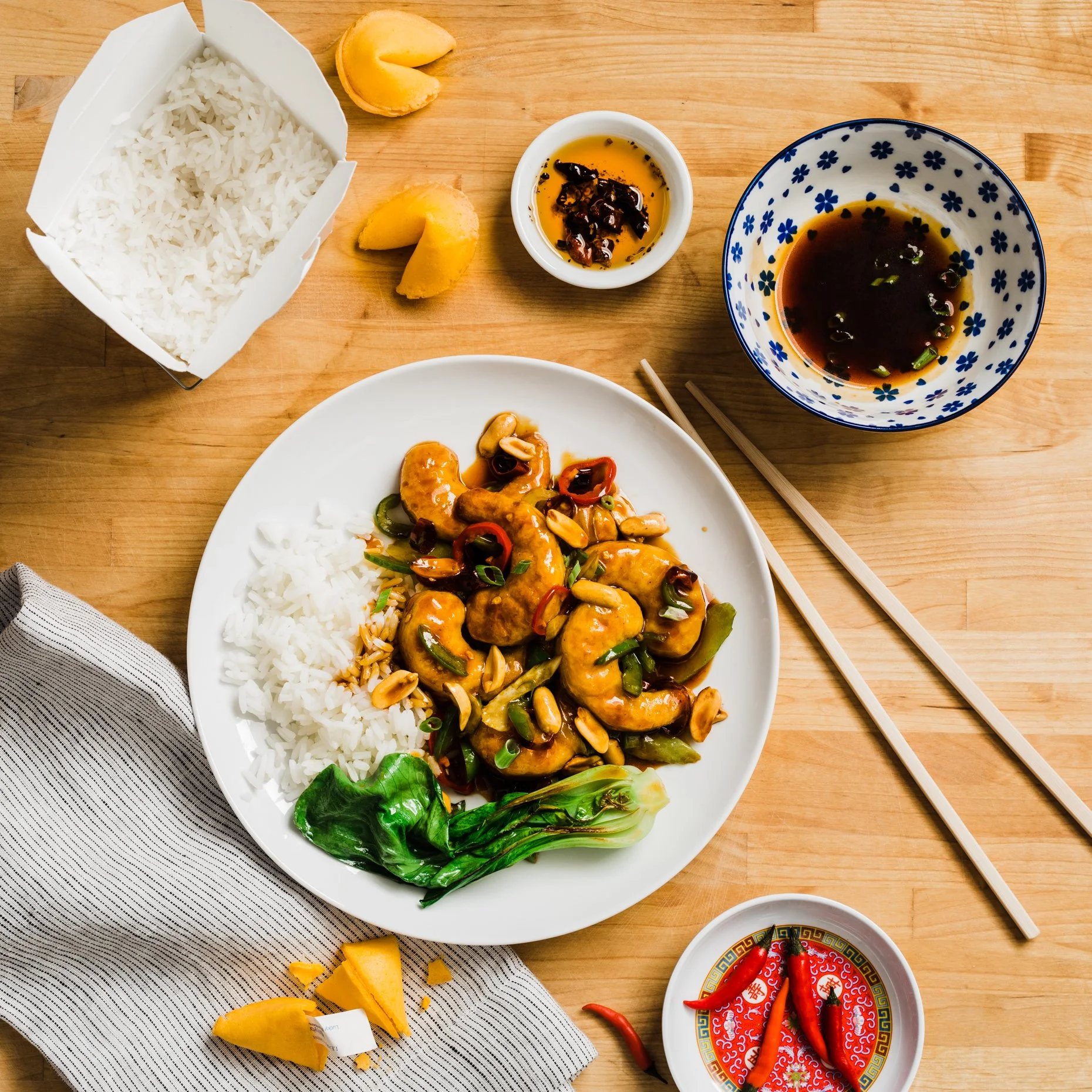 A plate with stir-fried chicken with vegetables and rice, accompanied by soy sauce and chili oil, on a wooden table with chopsticks, chili peppers, and pickled vegetables.