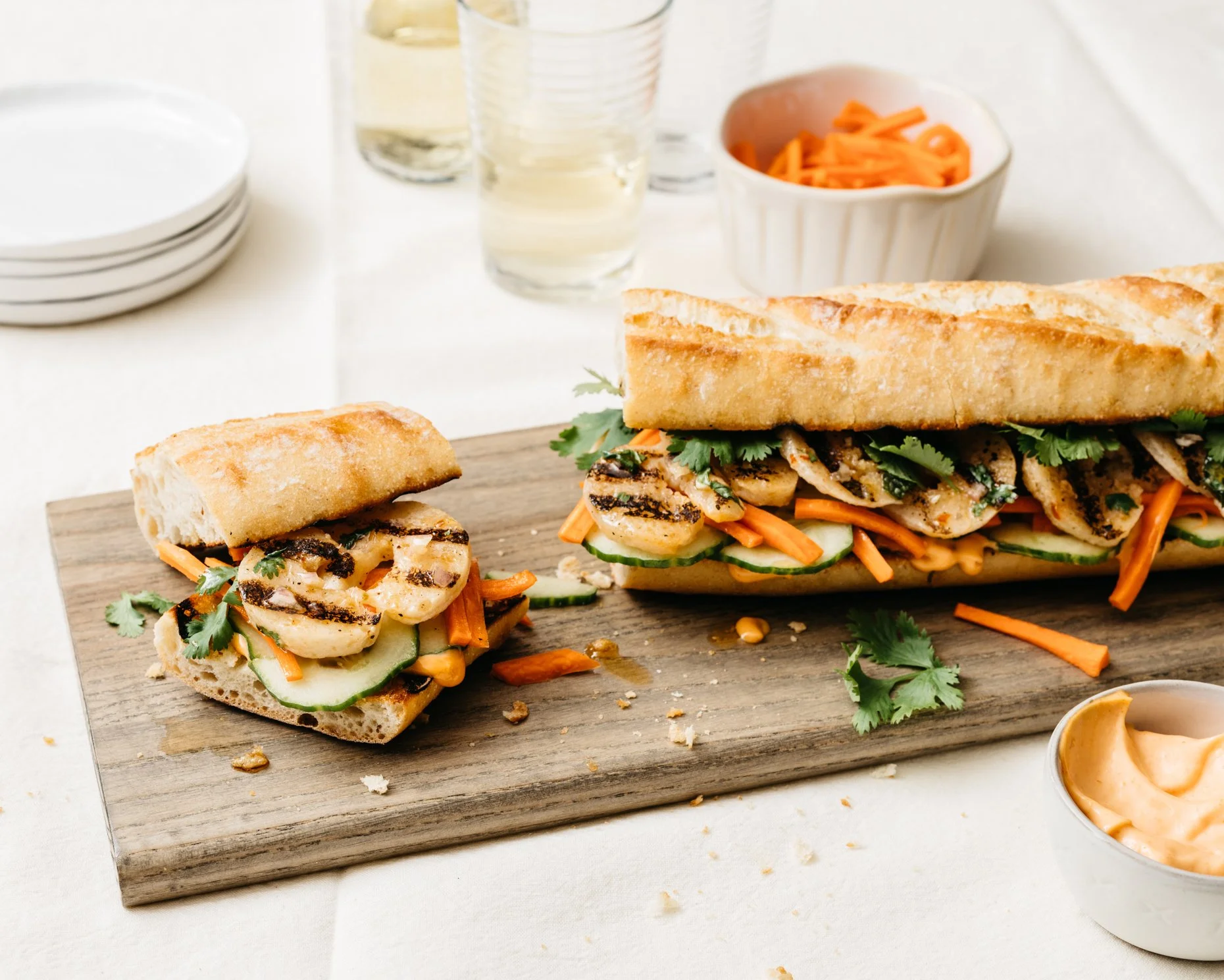 Grilled plant-based shrimp sandwich with vegetables and sauce on a wooden board, with glasses and small dishes in the background.