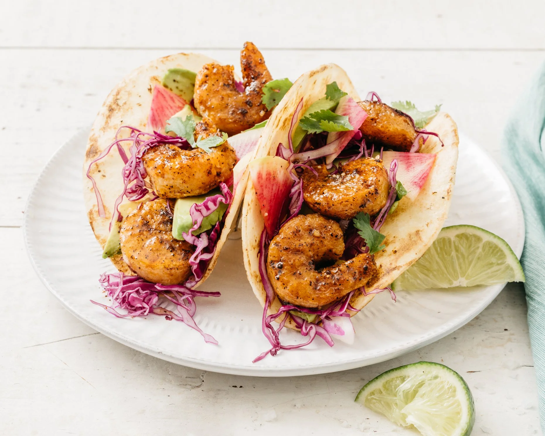 Two plant-based shrimp tacos on a white plate with sliced lime on the side.