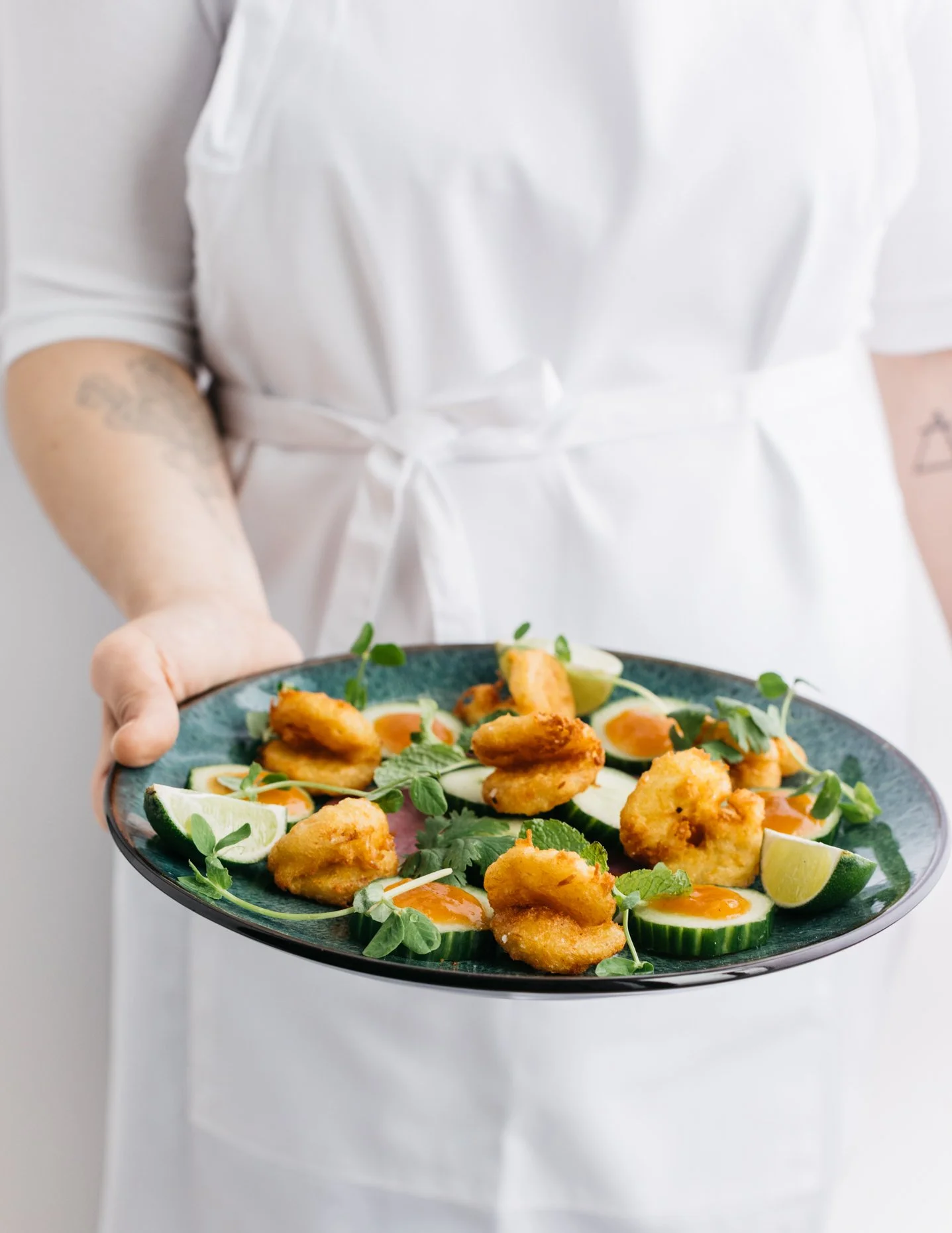 Person holding a plate of fried plant-based shrimp garnished with cucumber slices, lime wedges, and  microgreens.