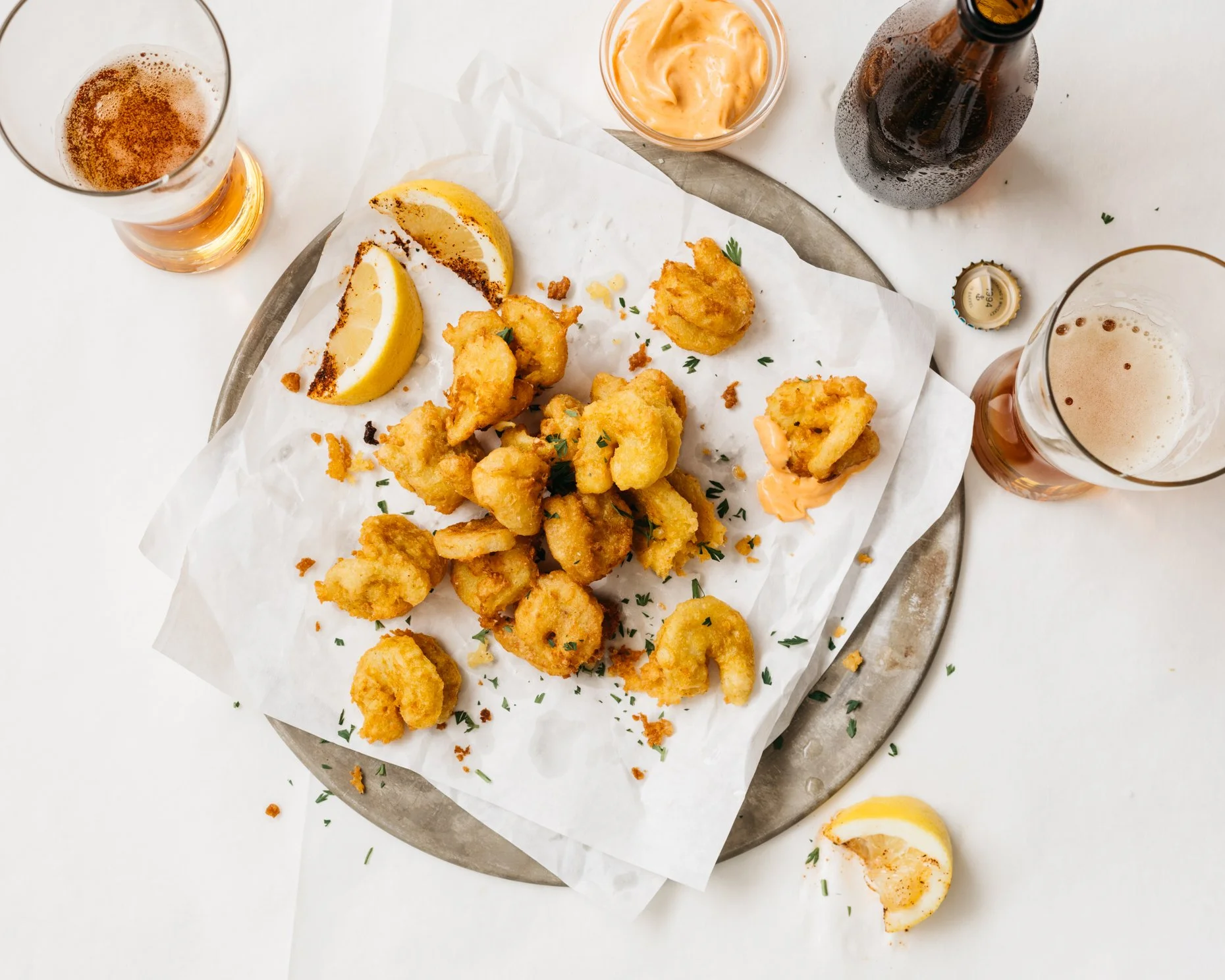Fried plant-based shrimp with lemon wedges on a silver platter, accompanied by two glasses of beer, a bottle of beer, and a small bowl of dipping sauce.