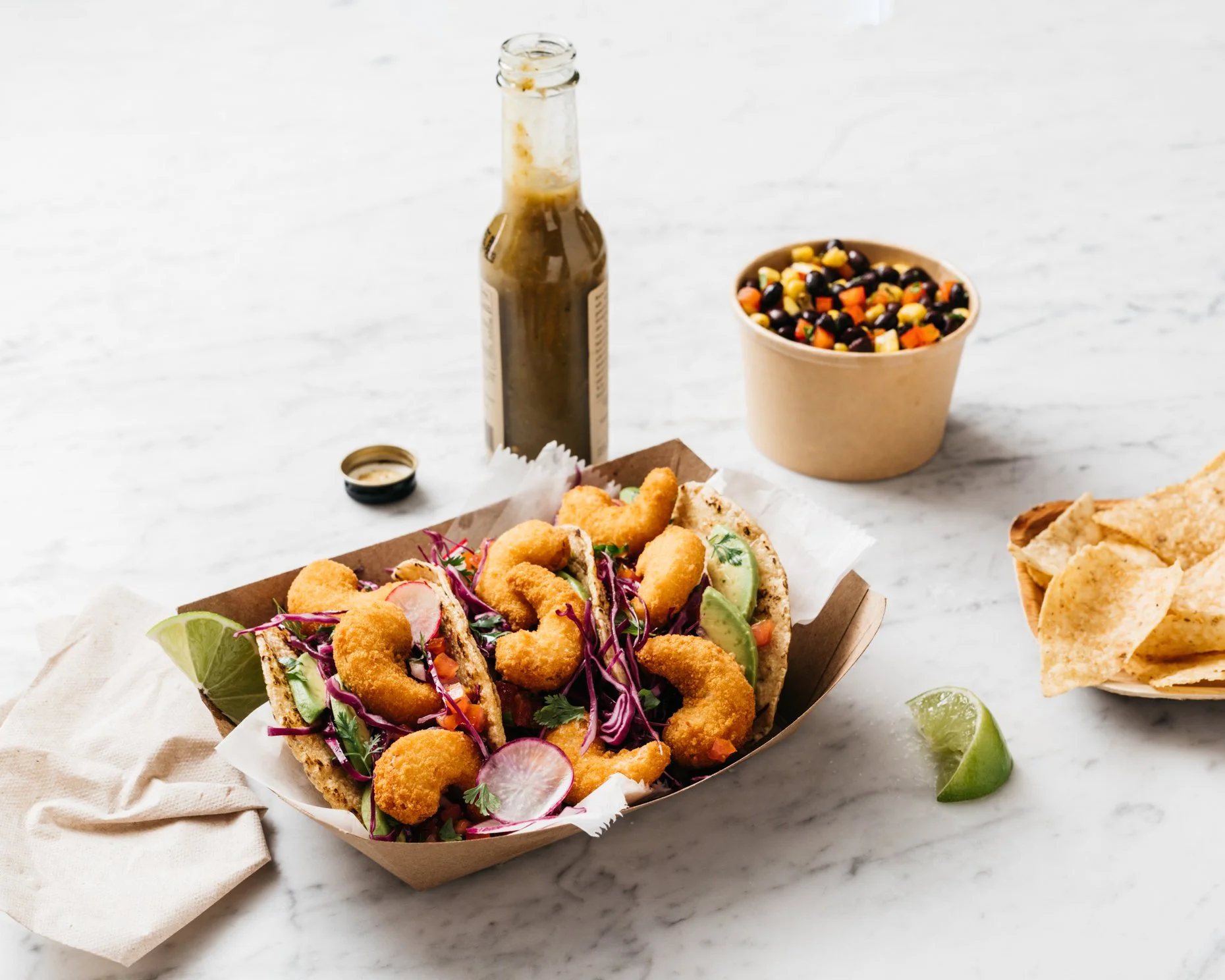 plant-based fish tacos with fried fish, cabbage slaw, and avocado slices, served in a paper tray on a white marble surface. There is a bowl of black bean salad, a bottle of sauce, tortilla chips with lime wedge, and a napkin nearby.