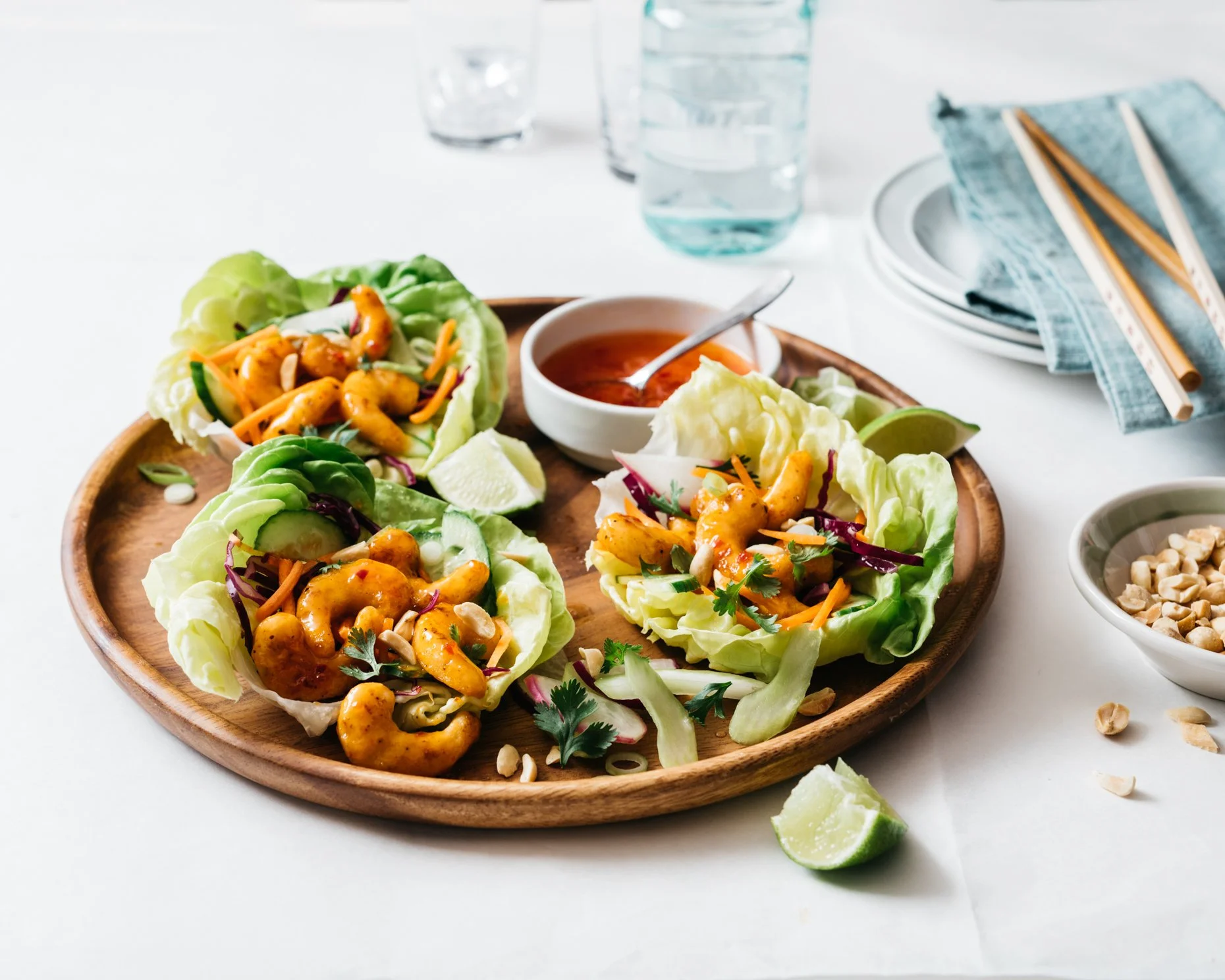 Three lettuce wraps with plant-based shrimp, vegetables, and sauce on a wooden platter, with lime wedges, a bowl of chopped peanuts, and a small bowl of sauce on a white table.