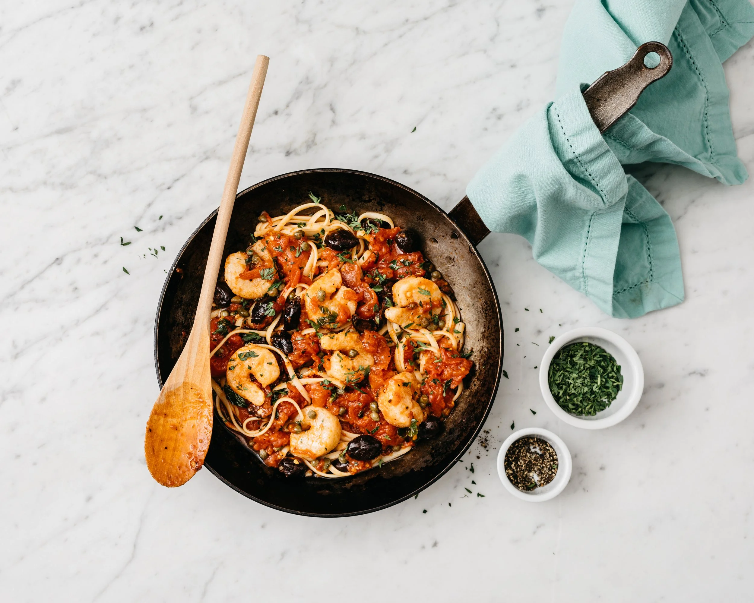 A skillet of plant-based shrimp and spaghetti with tomato sauce garnished with herbs on a white marble surface with small bowls of chopped herbs and black pepper, and a wooden spoon