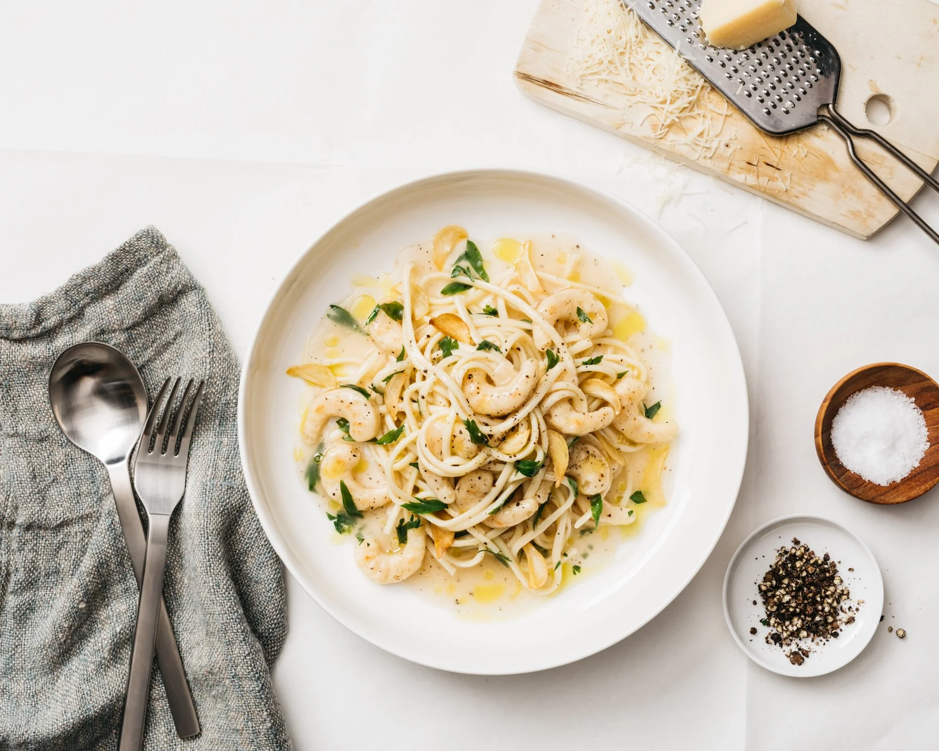 Plate of plant-based shrimp scampi pasta with garlic, herbs, and olive oil on a white table, with a spoon and fork on a gray napkin, grated cheese, salt, and black pepper on the side, and a cheese grater in the background.