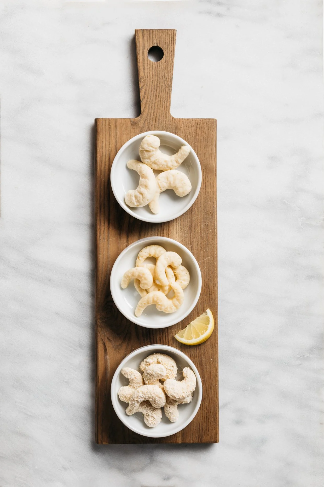 Three white bowls with shrimp on a wooden board, lemon wedge, on a white marble surface.