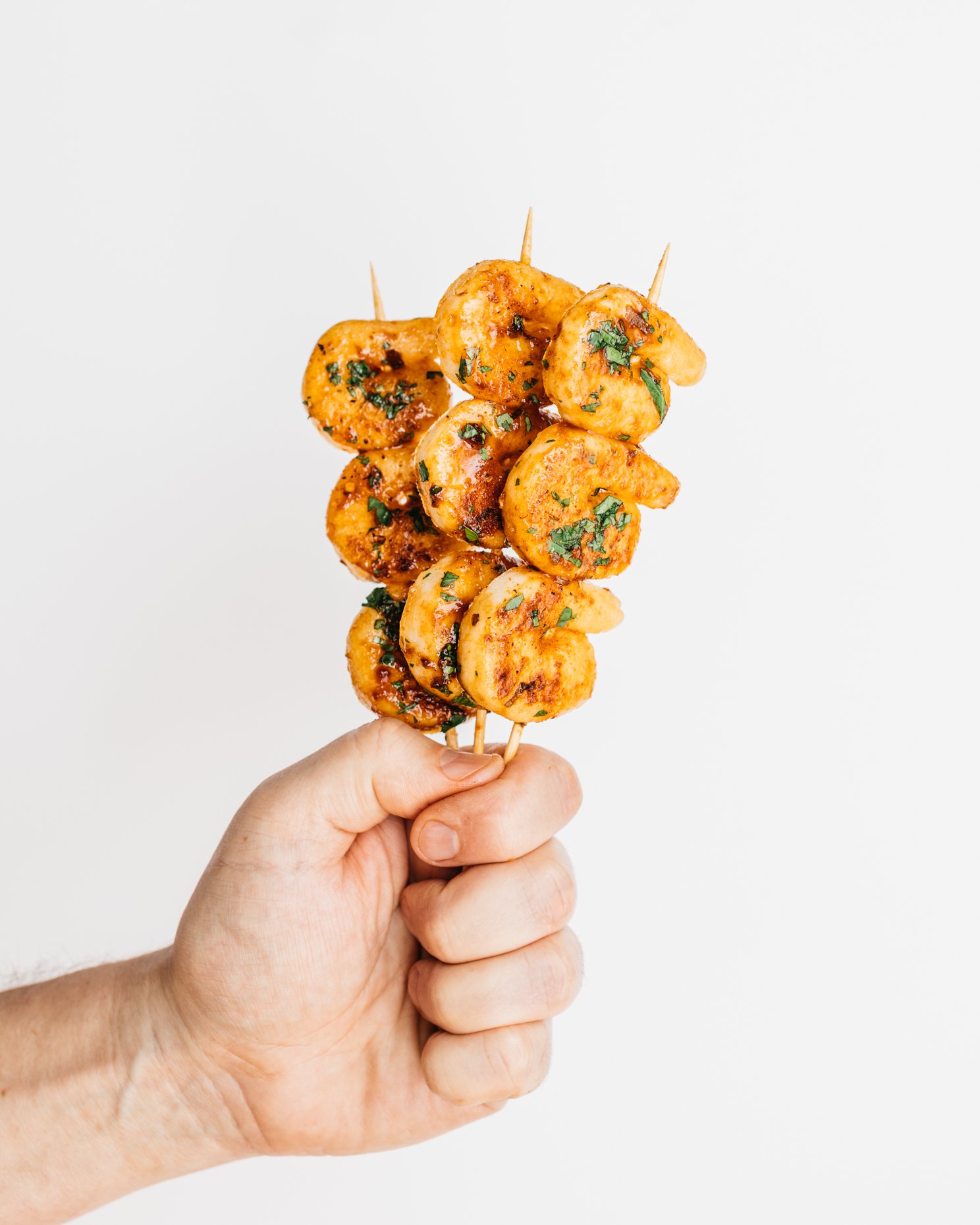 Hand holding a skewer with grilled plant-based shrimp garnished with chopped herbs on a white background.