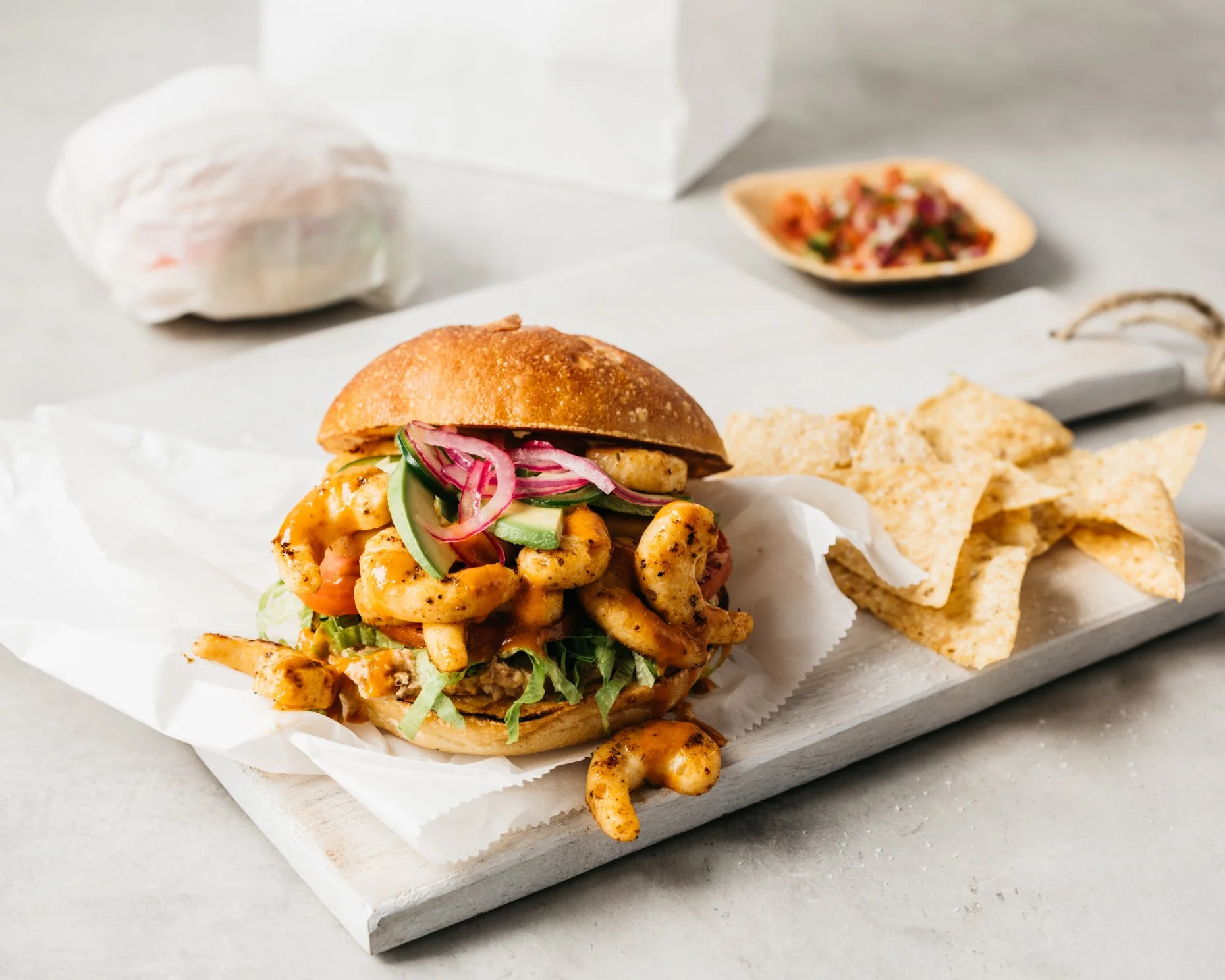 A plant-based shrimp po' boy sandwich with pickles, onions, and lettuce on a bun, served with tortilla chips and a small side of salsa.