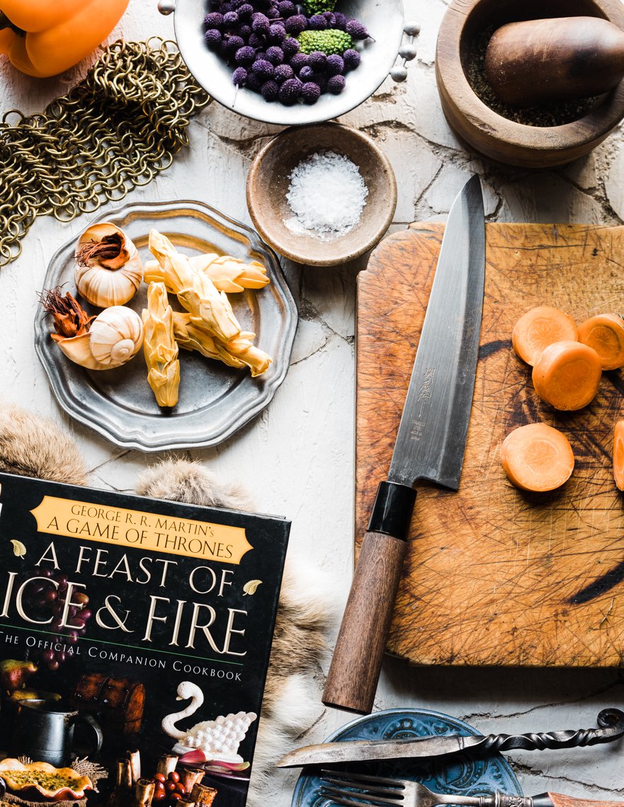 A chopping board with sliced carrots, a kitchen knife, a cookbook titled "A Feast of Ice and Fire," garlic, salt, and various bowls containing ingredients, around a rustic white surface.