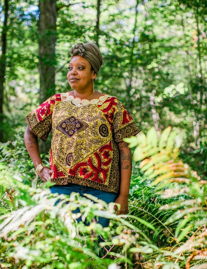 A woman standing outdoors in a green forest, wearing a colorful patterned shirt and a headwrap, with her hand on her hip, surrounded by lush greenery.