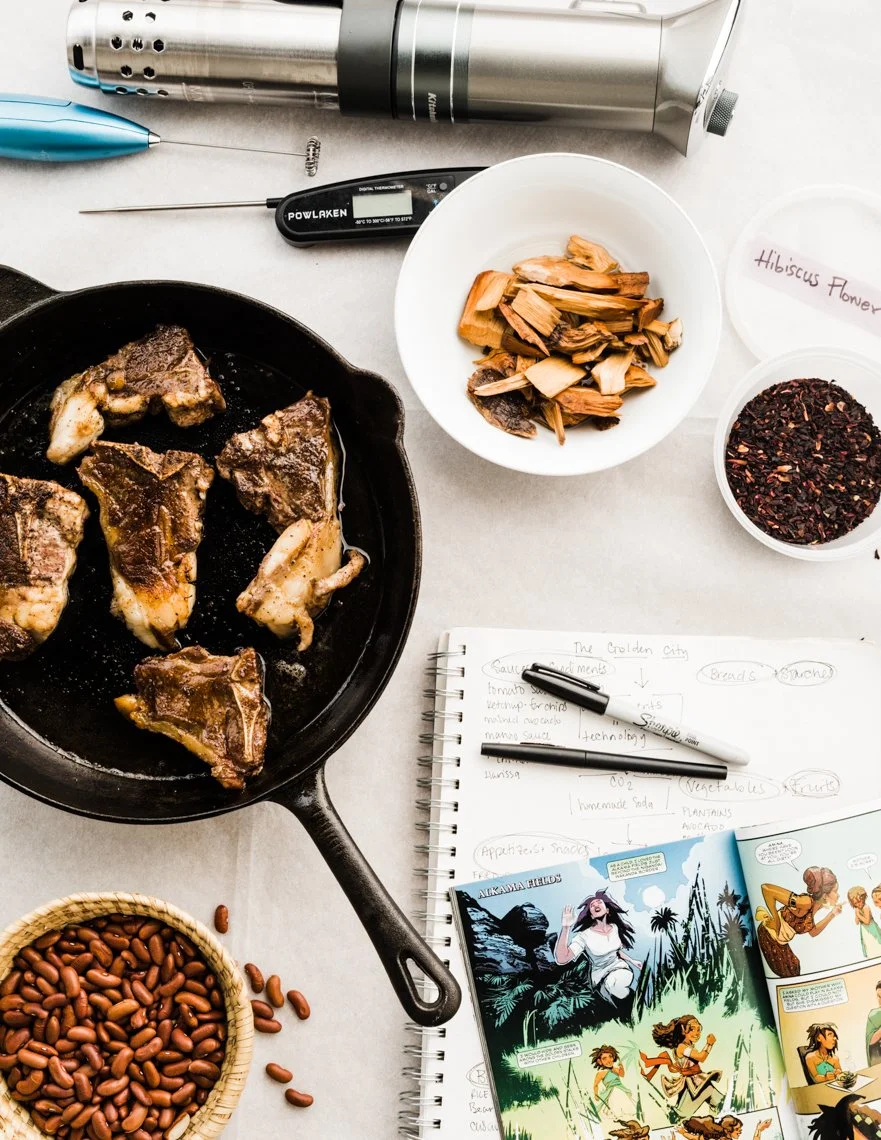 Cooking scene with raw meat in a cast iron skillet, dried mushrooms in a bowl, roasted beans, a thermometer, a notebook with notes, a pen, a comic book, and herbal tea in containers.