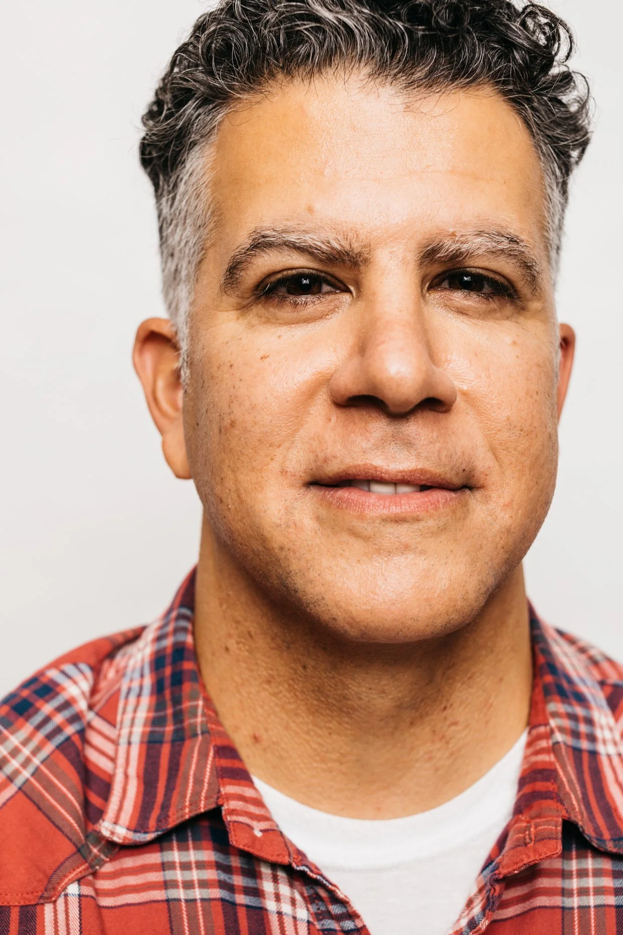 Close-up of a middle-aged man with short, dark and gray hair, wearing a red plaid shirt, smiling slightly against a plain white background.