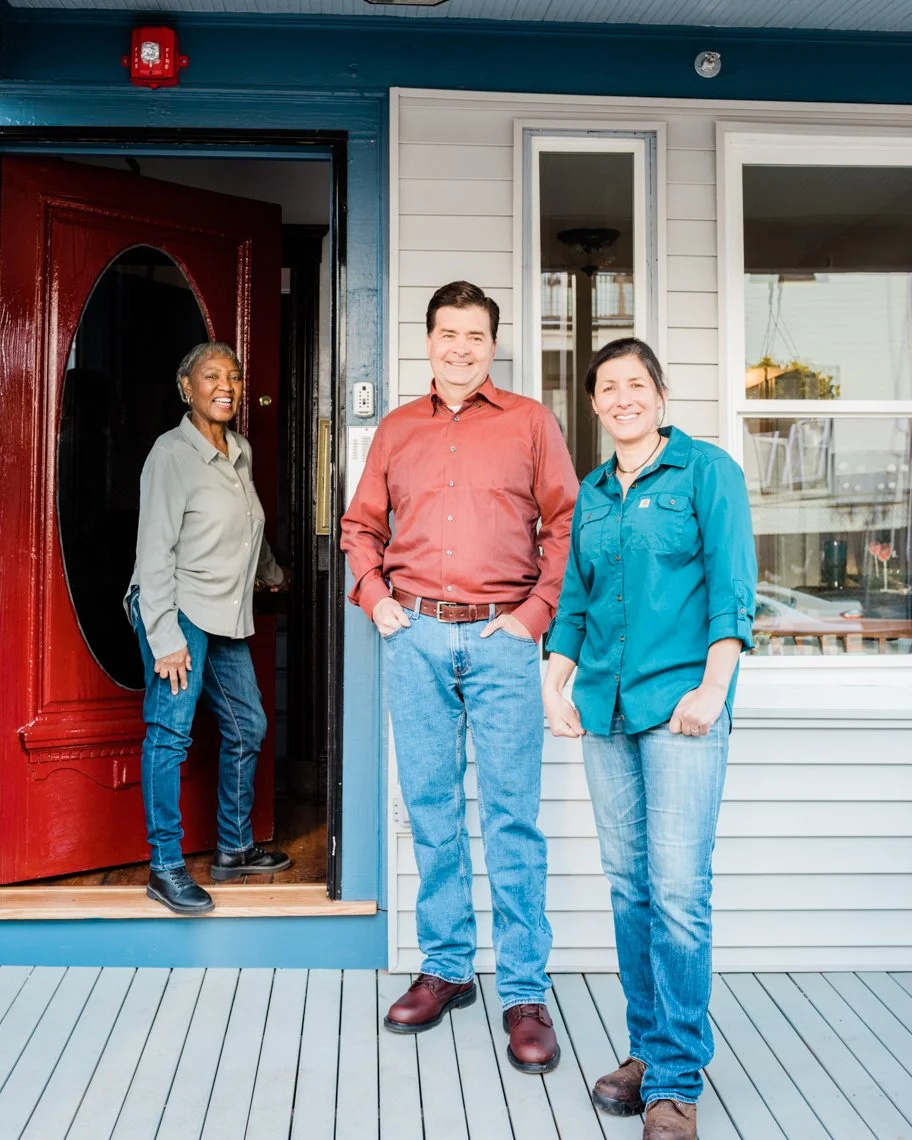 Three people standing outside a house, smiling and posing for the photo. A woman is standing at the door, and a man and woman are standing on the porch.