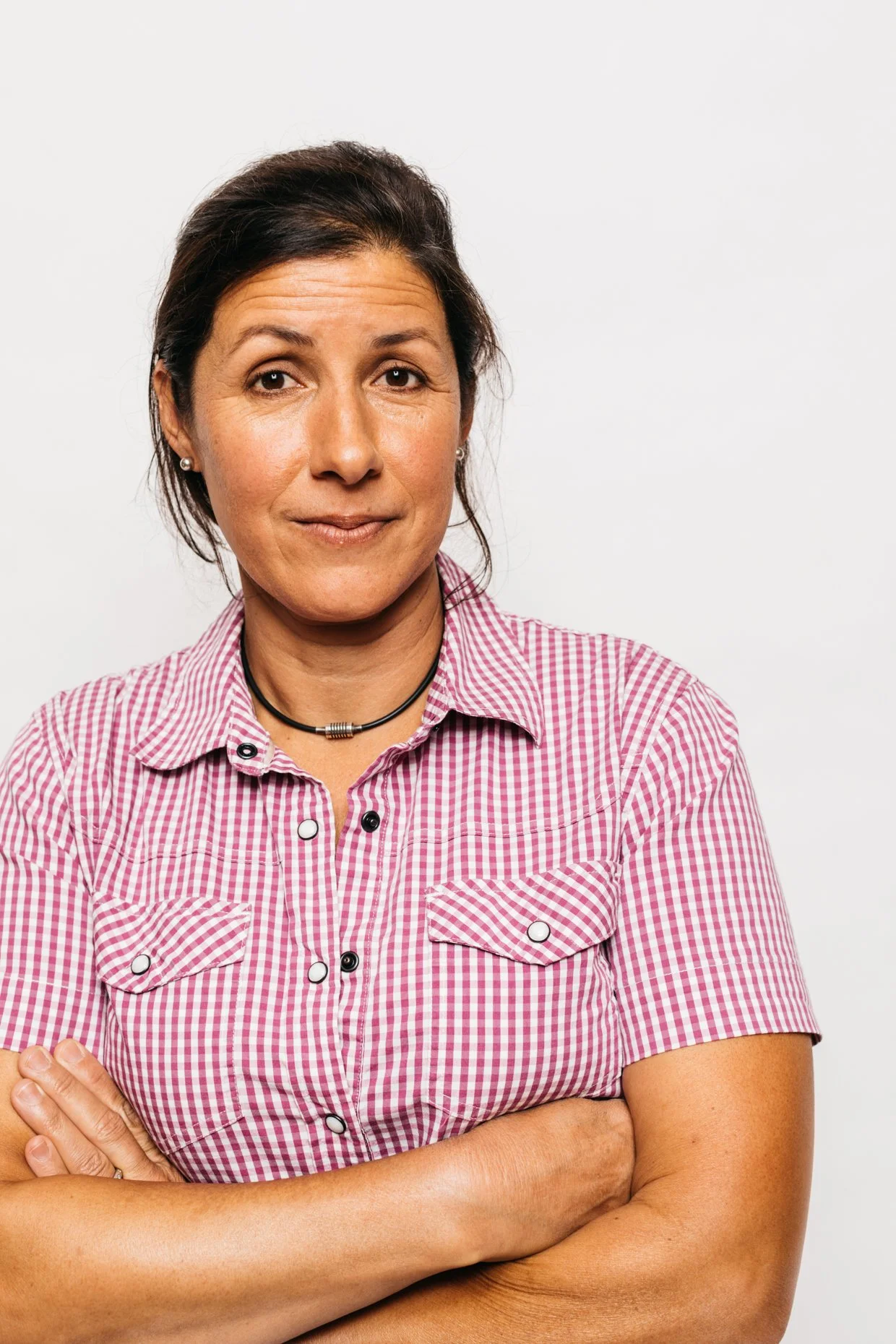 A woman with dark hair in a checkered pink and white shirt, crossed arms, and a confident expression, standing against a plain white background.