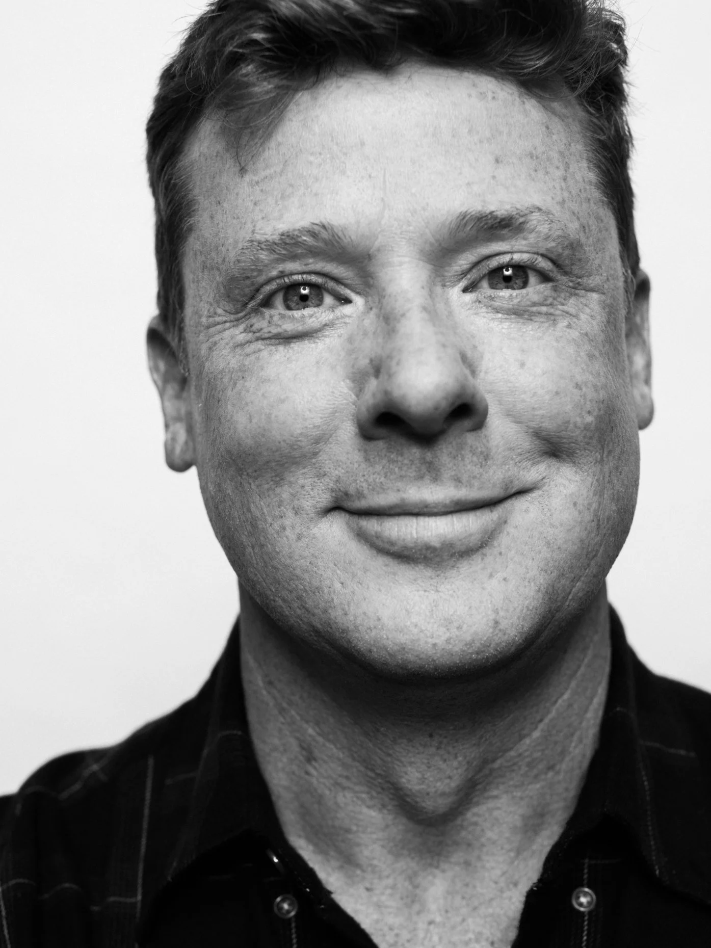 A smiling man with short hair and light skin, wearing a dark shirt with a checkered pattern, in black and white.
