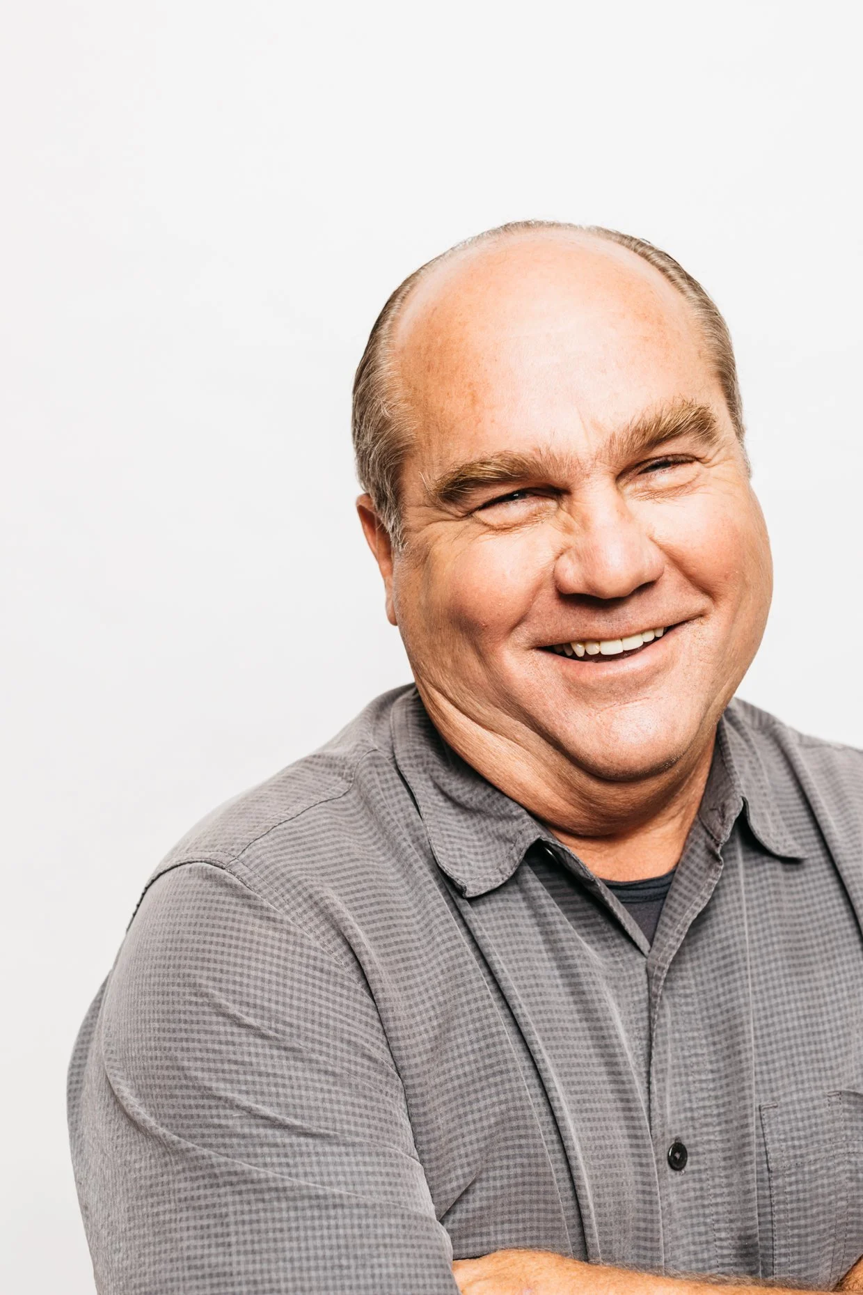 A smiling middle-aged man with light skin, receding hair, and wearing a gray button-up shirt, caught mid-laugh against a plain white background.
