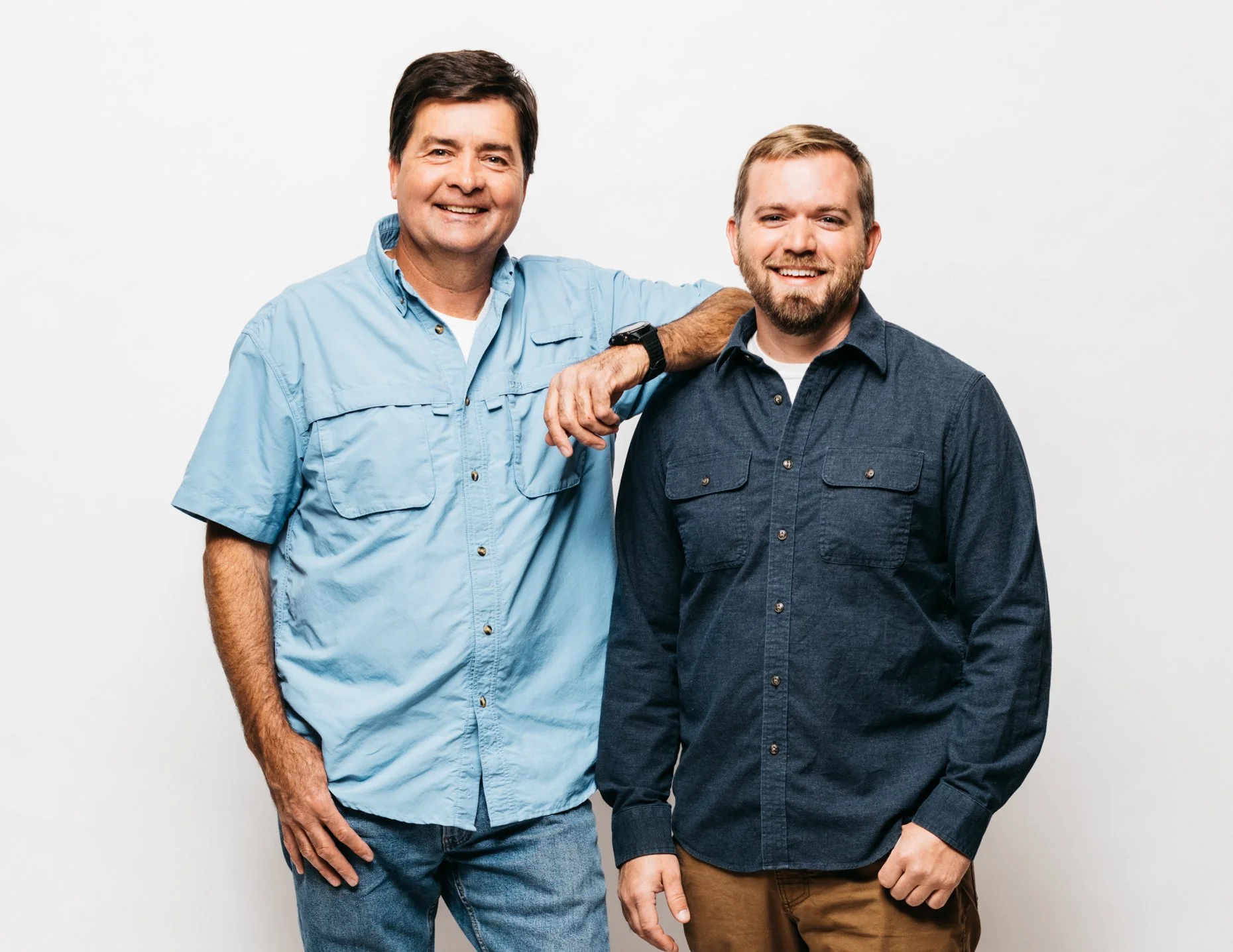 Two smiling men standing against a plain white background, one with his arm resting on the other's shoulder, wearing casual shirts.