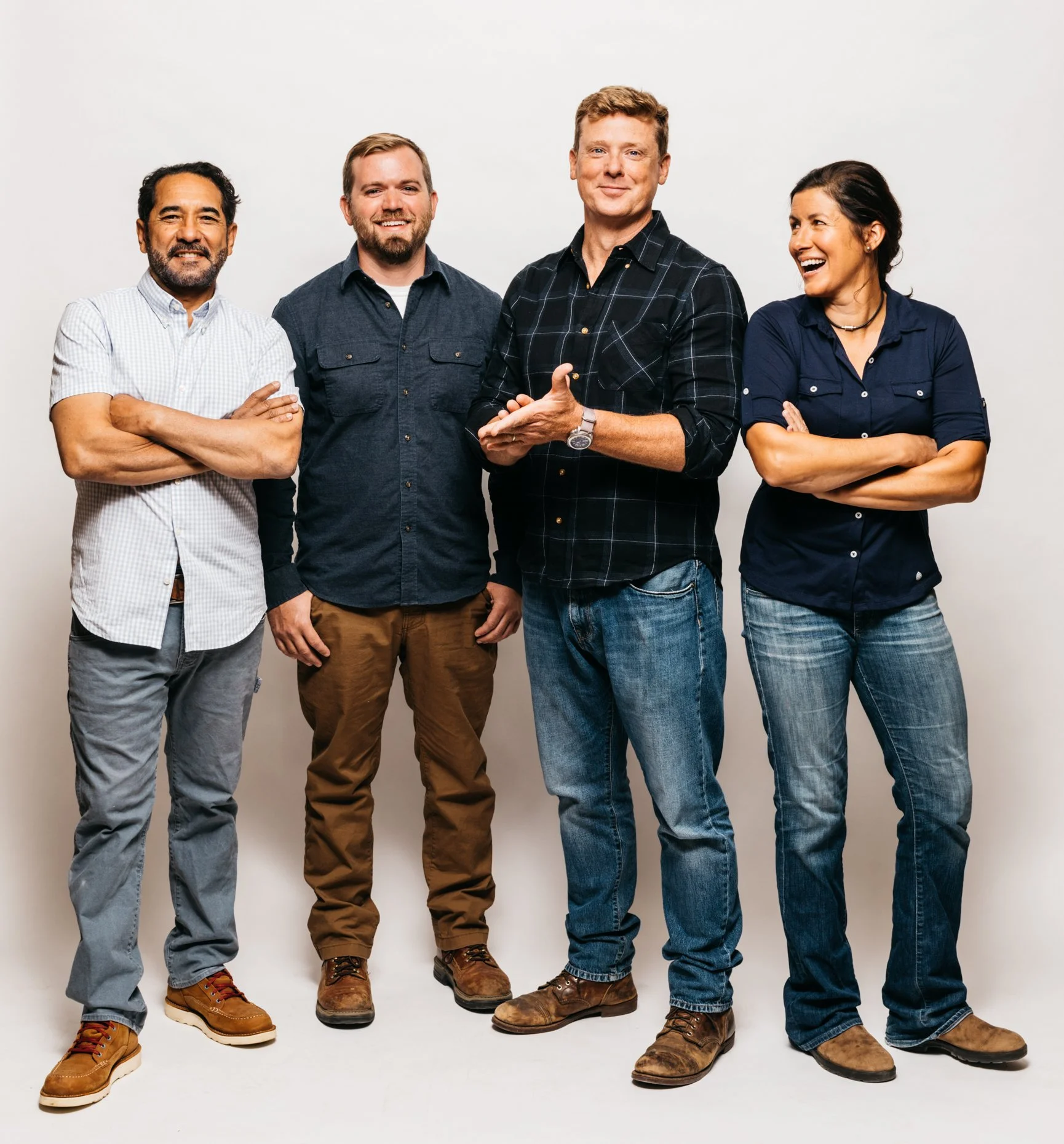 Four adults standing together against a blank wall, smiling and laughing, dressed casually in jeans and button-up shirts.
